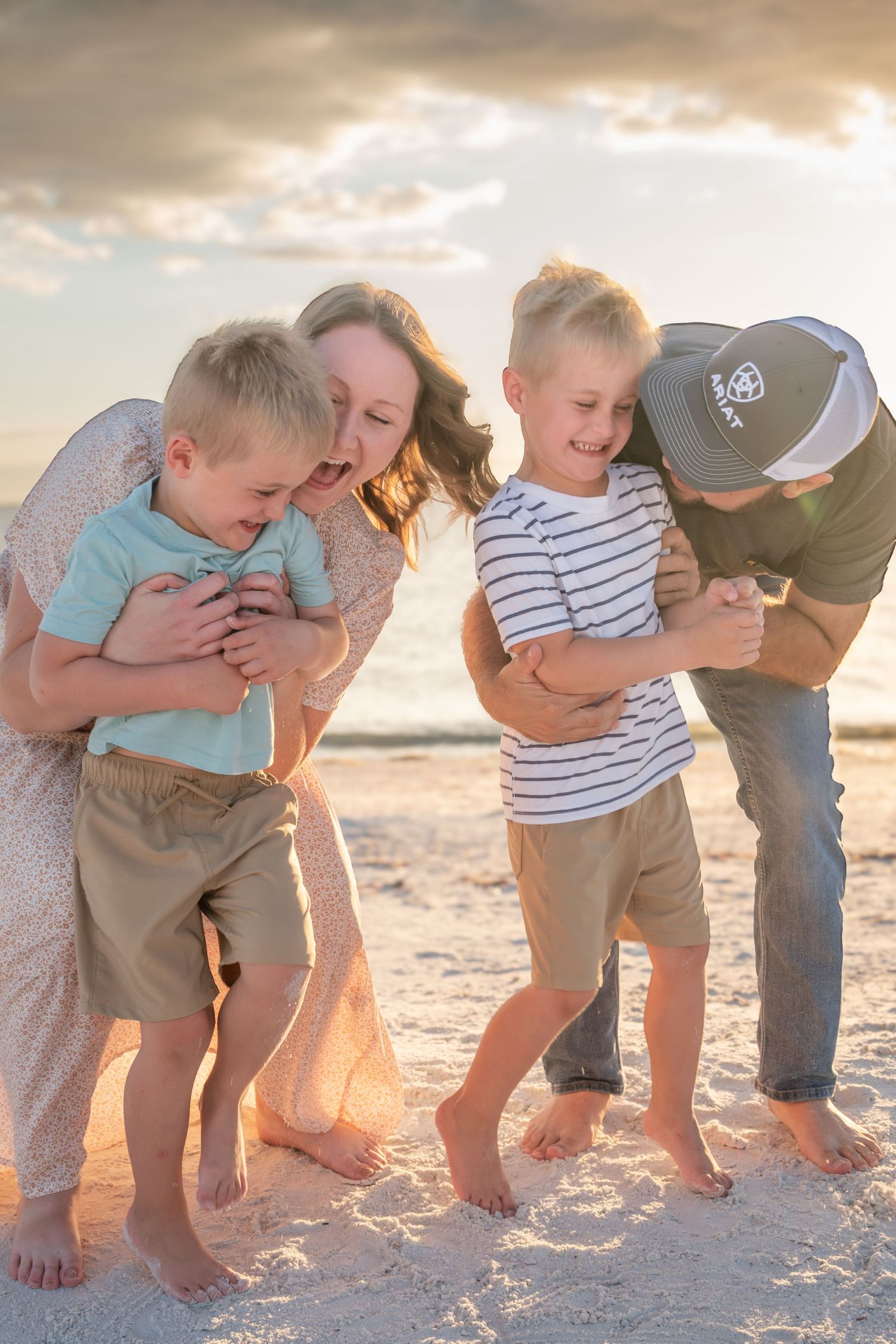 Family of four playfully hugging two young boys on sandy beach during golden sunset.