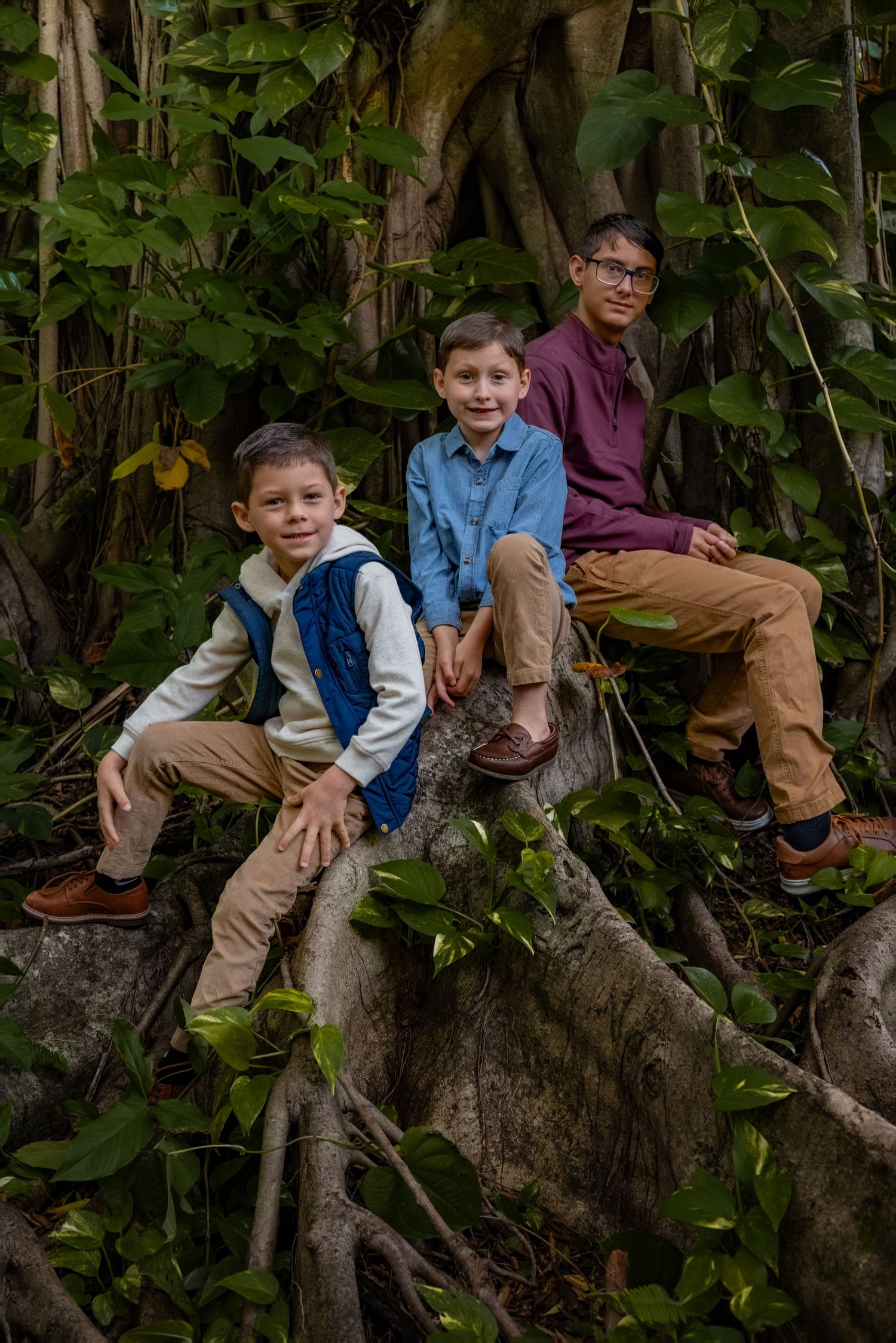 Three boys pose on large tree roots surrounded by lush tropical greenery in a forest setting.