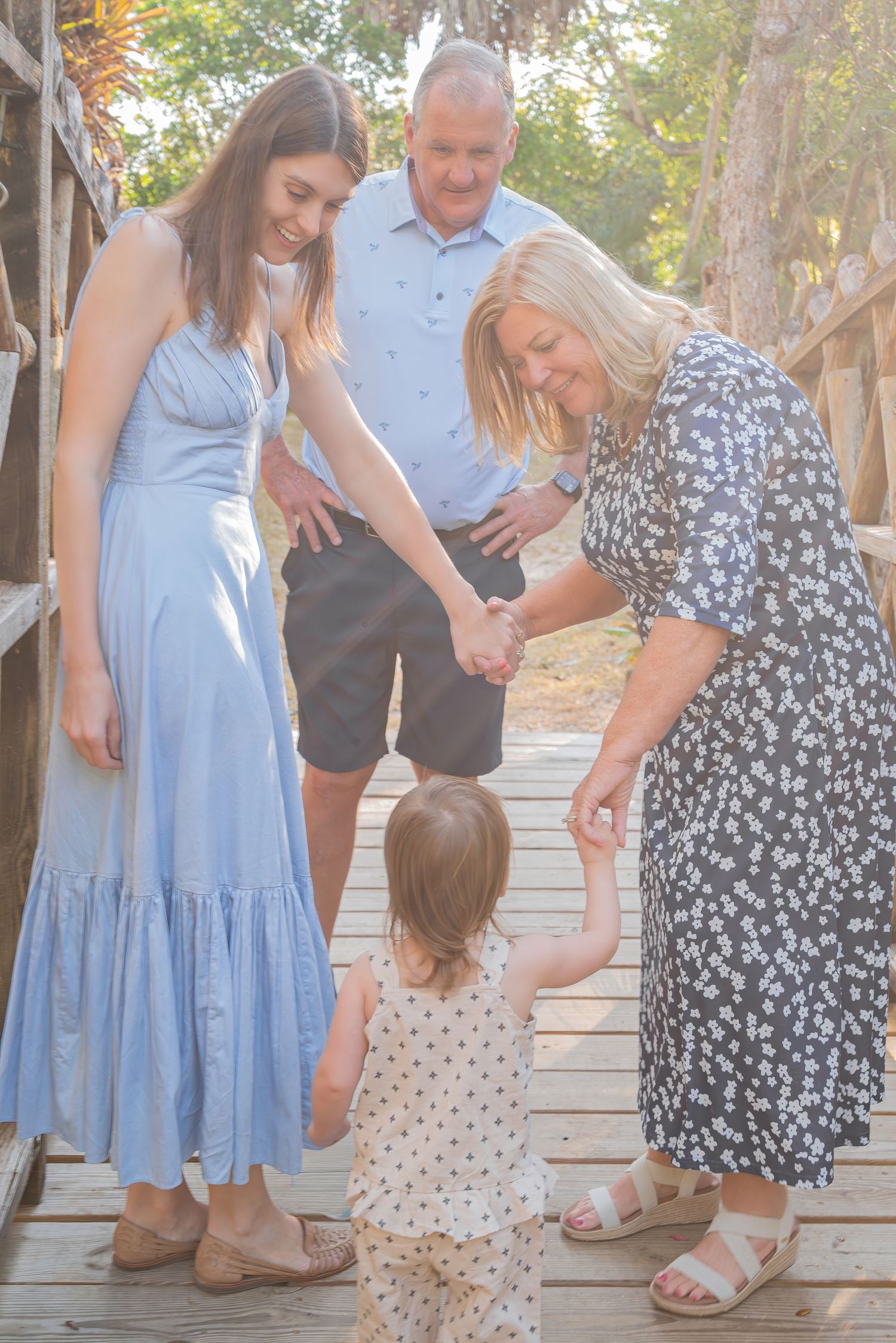 Three adults interact warmly with a toddler on a wooden deck surrounded by trees.