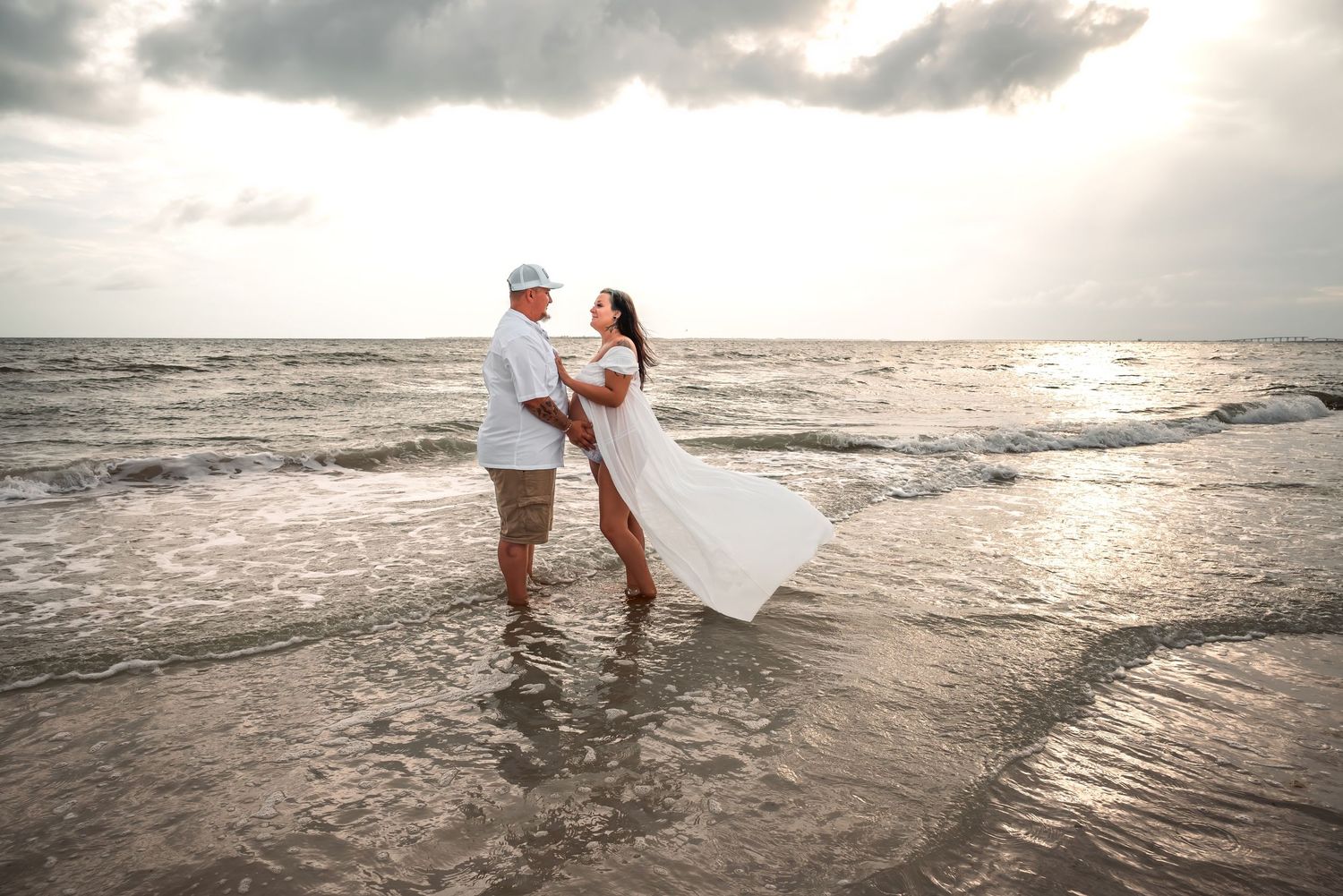 Couple in white attire holding hands on a beach at sunset, waves washing over their feet.