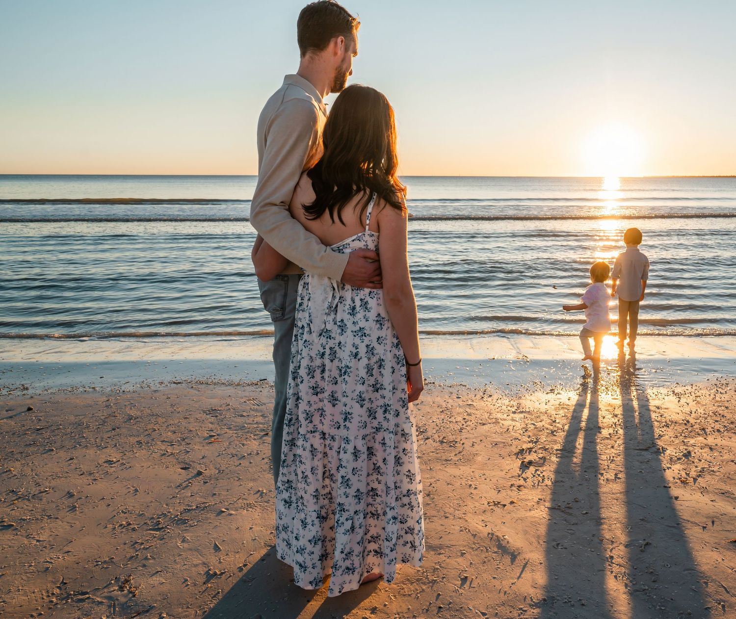 Romantic couple embracing on beach at sunset with children nearby.