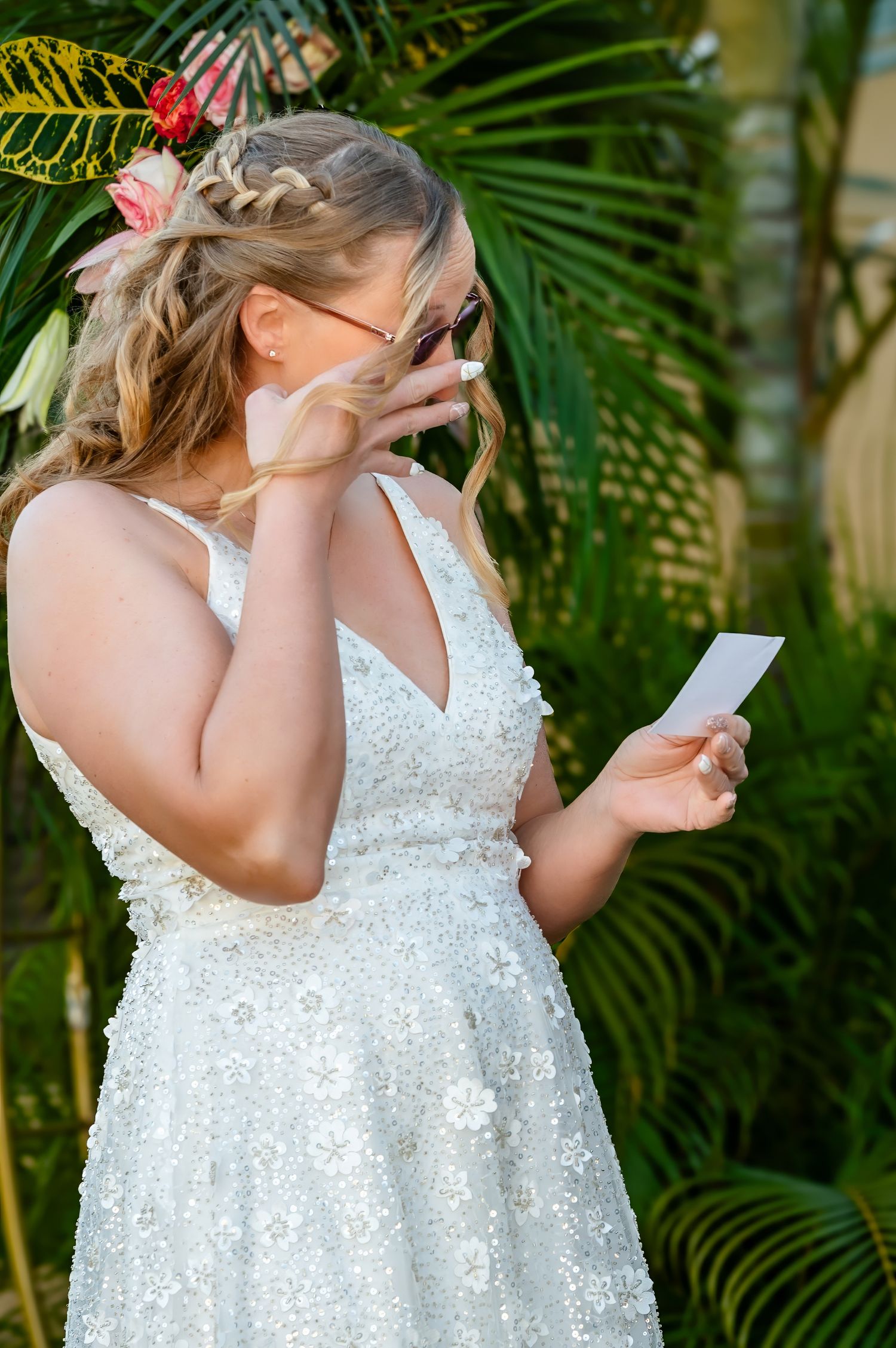 Emotional bride in a white lace gown reading vows while wiping tears, surrounded by tropical palms.