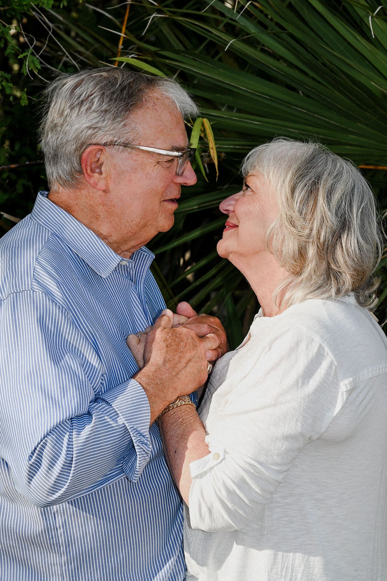 Senior couple holding hands and dancing together outdoors, smiling at each other with palm trees in the background.