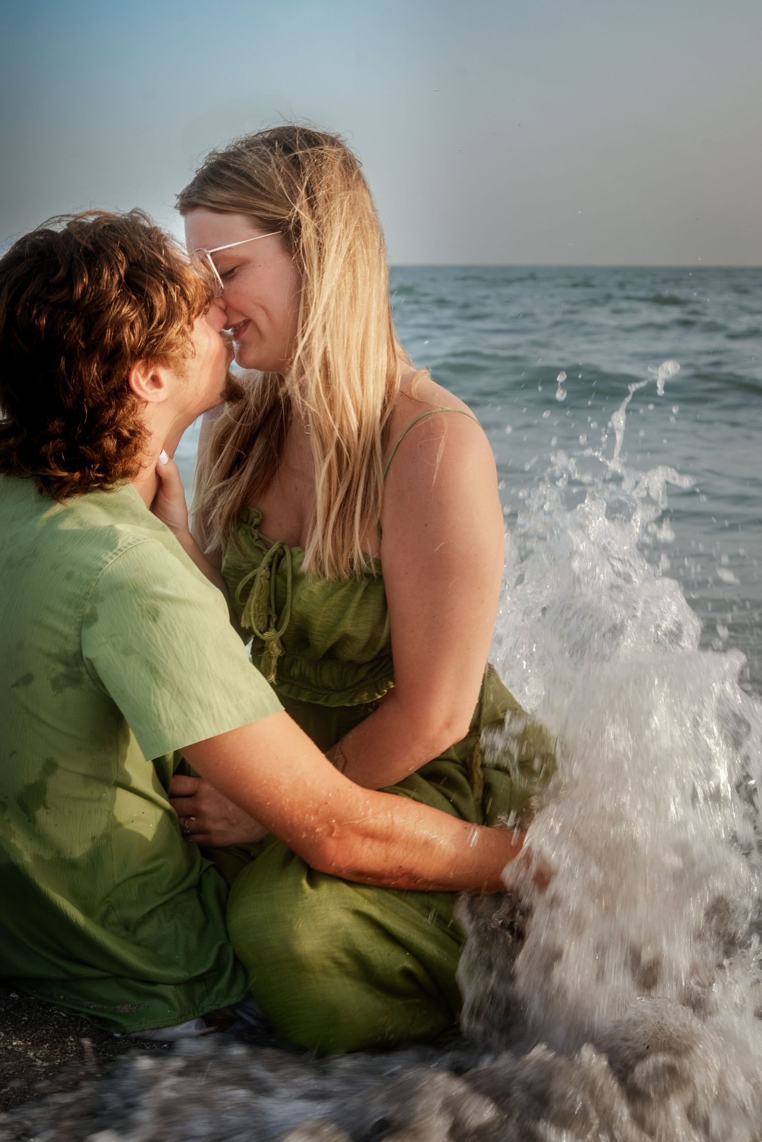 Couple sharing a kiss while sitting in ocean waves on beach, with water splashing around them at sunset.