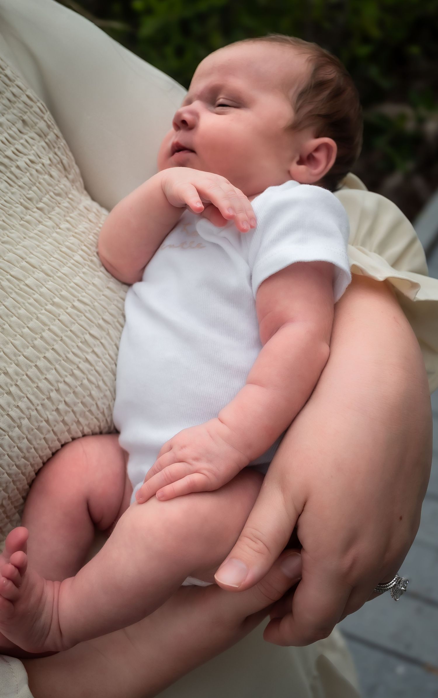 Newborn baby sleeping peacefully in white onesie cradled in mother's arms outdoors.