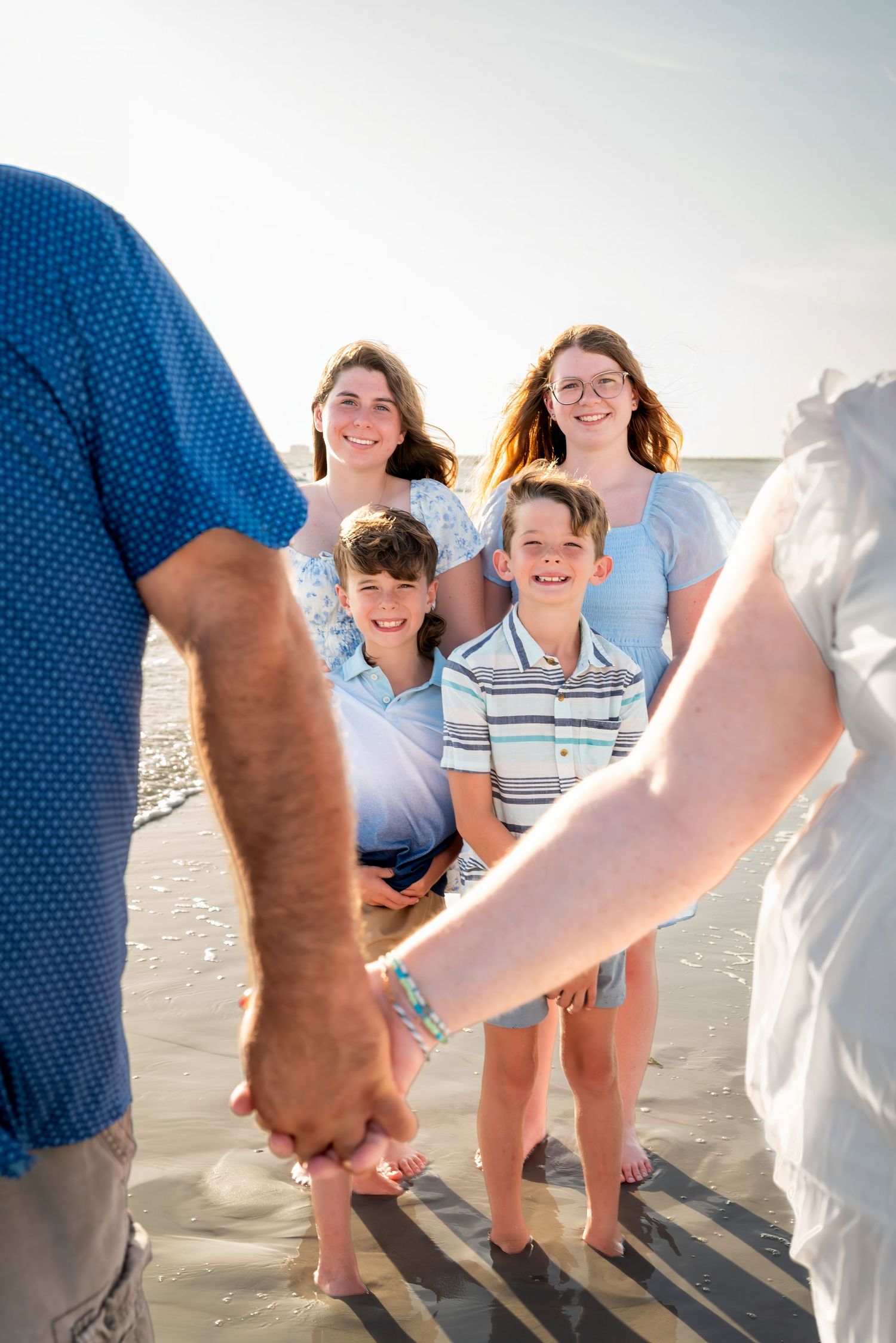Family of four children and parents holding hands on a sunny beach, smiling together near the ocean shoreline.