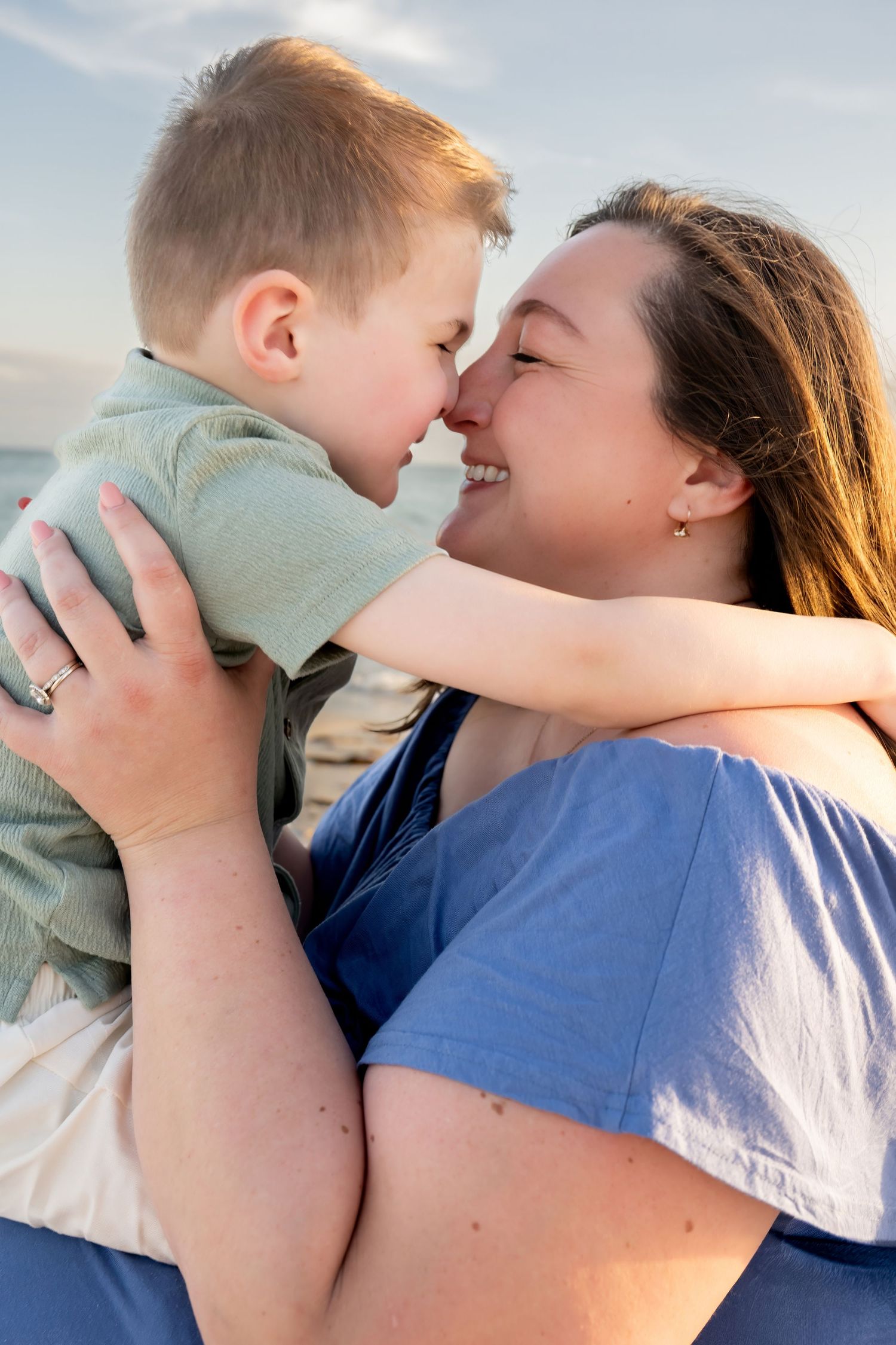 A smiling mother holds her young son close as they touch noses affectionately on a sunny beach.