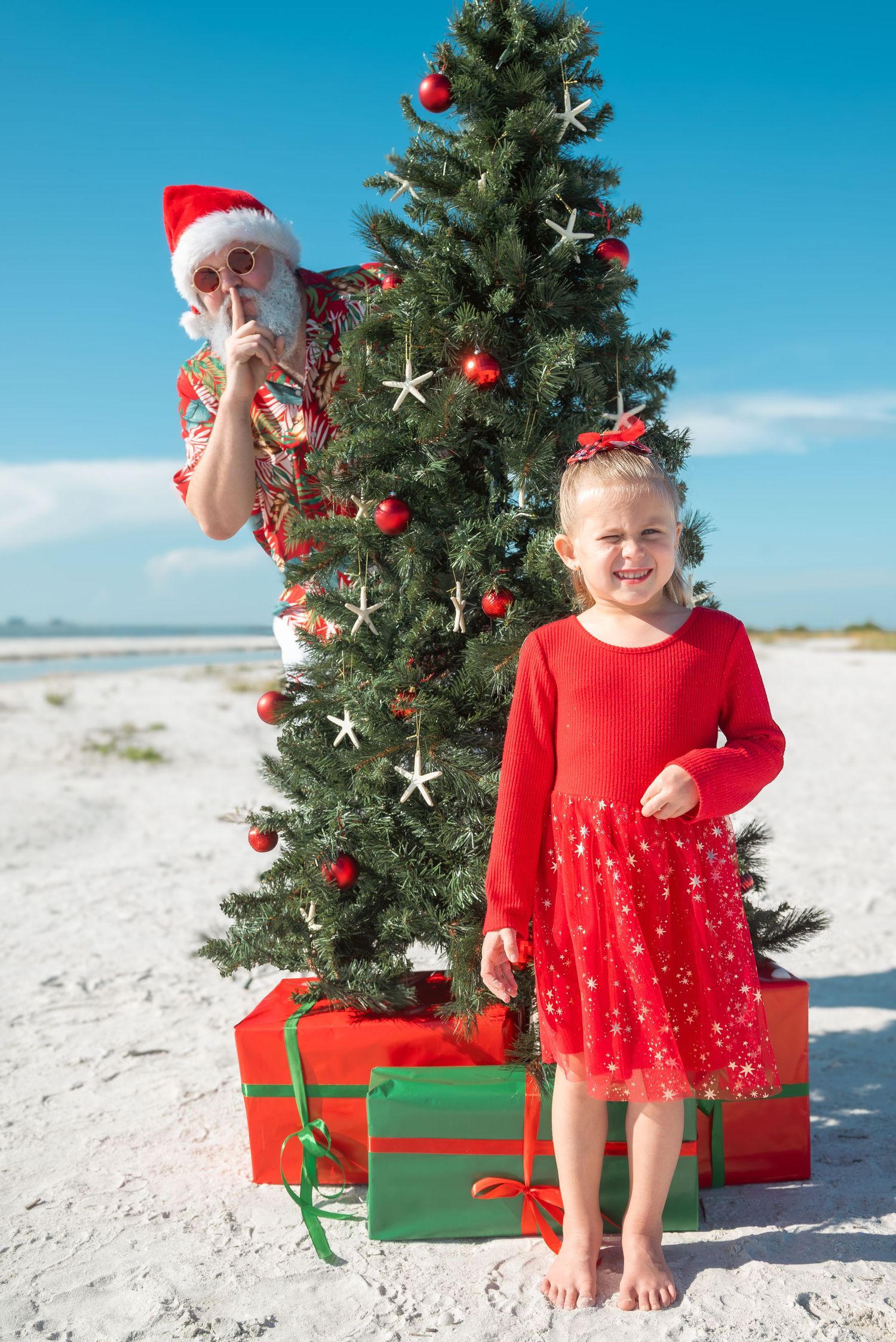 Little girl in red dress with Santa Claus hiding behind a beach Christmas tree.