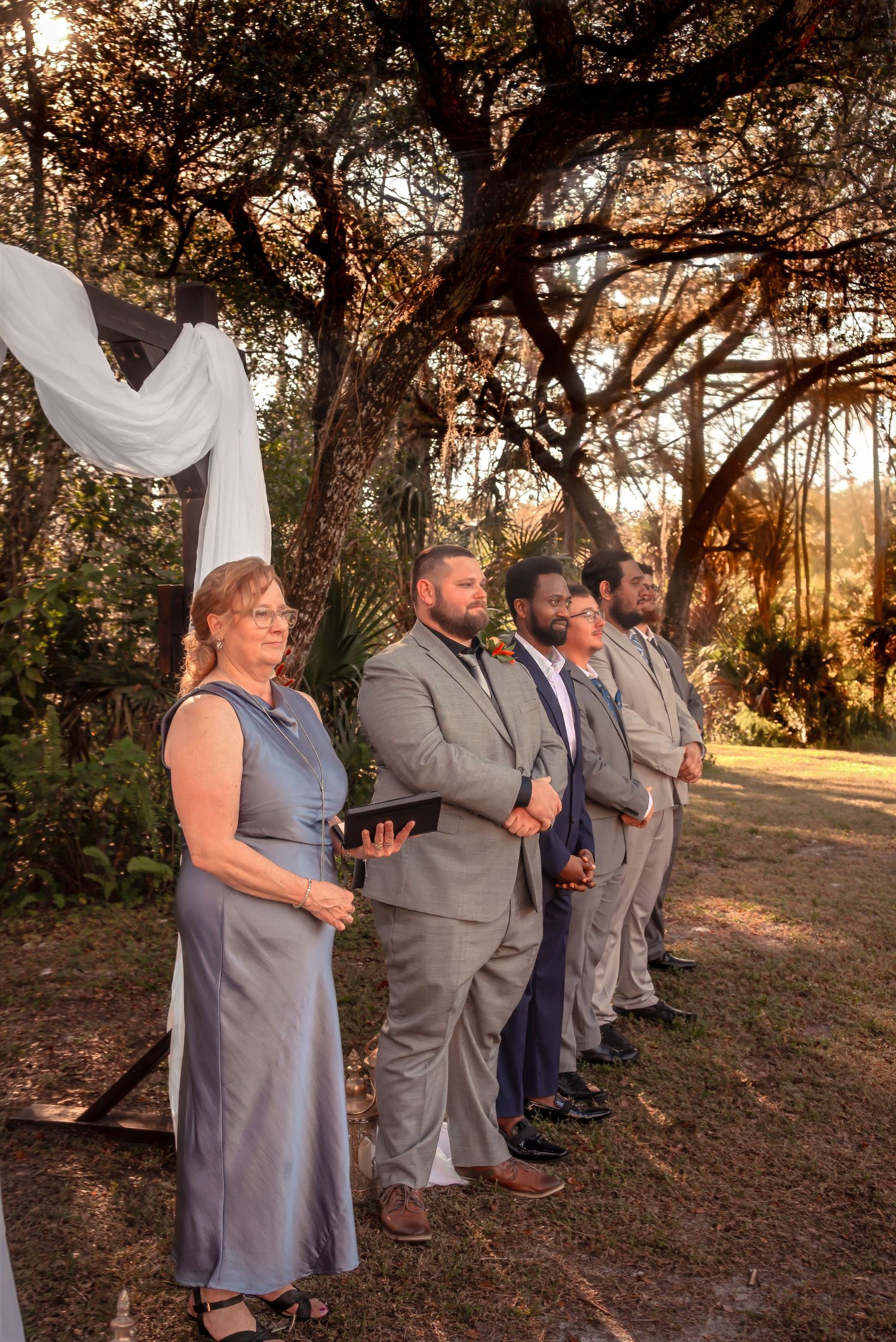 Wedding officiant and groomsmen stand beneath a draped ceremony arch during an outdoor sunset wedding.