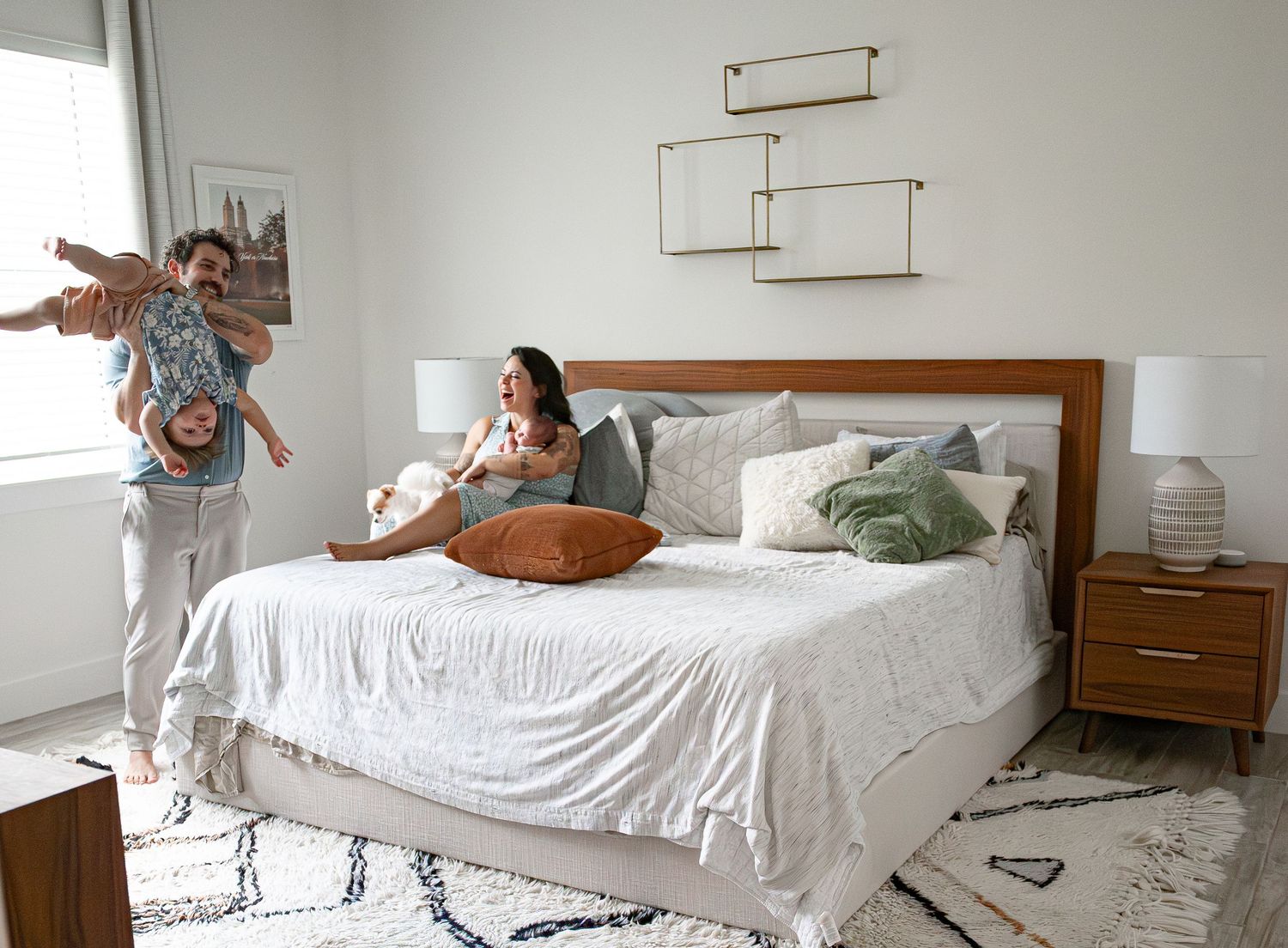 Joyful family in bright bedroom with dad lifting toddler and mom sitting on bed with newborn.