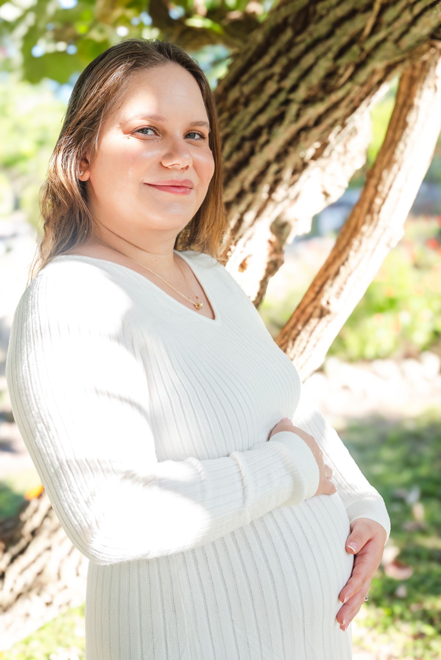 Smiling pregnant woman in white knit dress posing outdoors beside a tree in natural light.