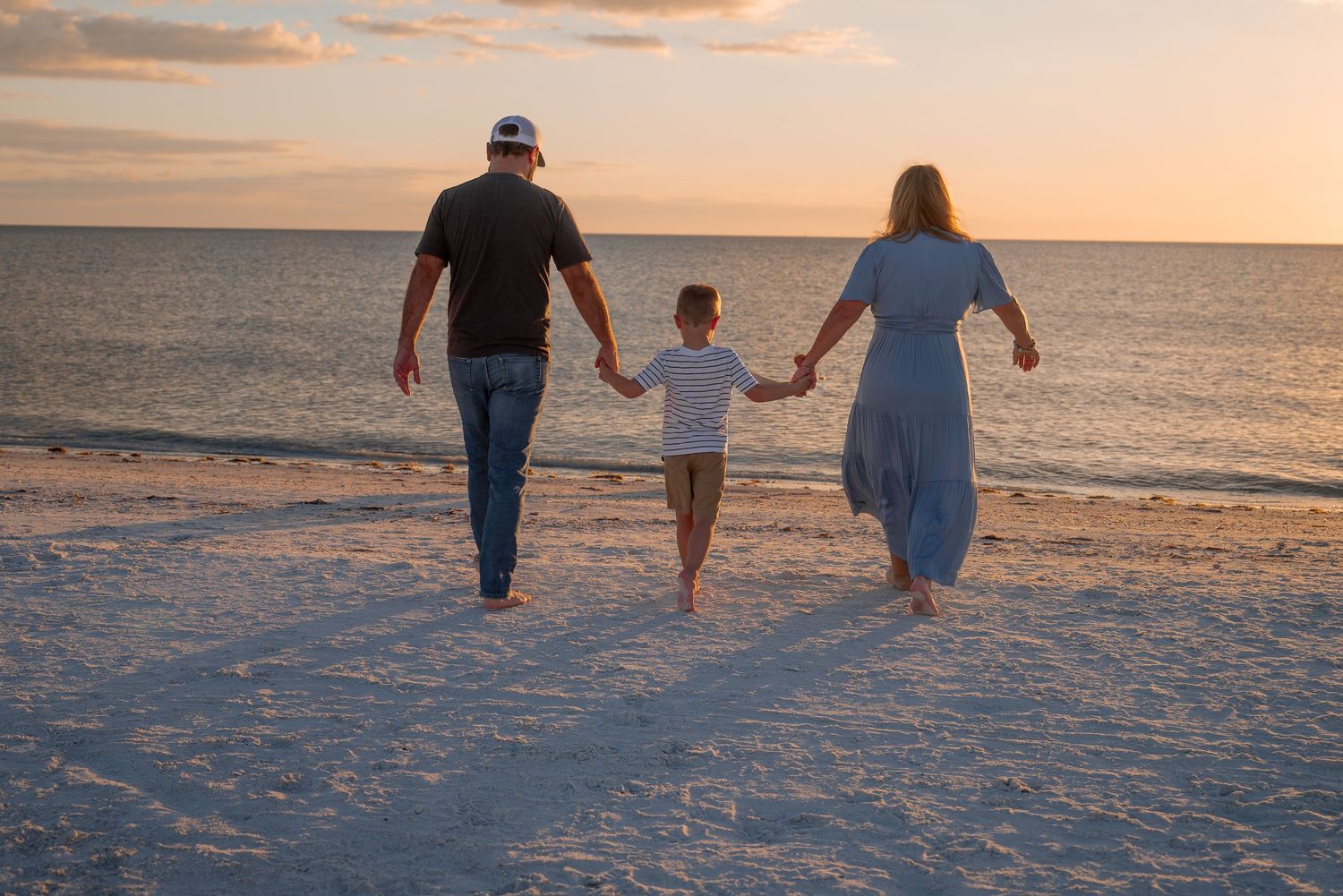 Family of three holding hands walking toward ocean at sunset on a white sandy beach.