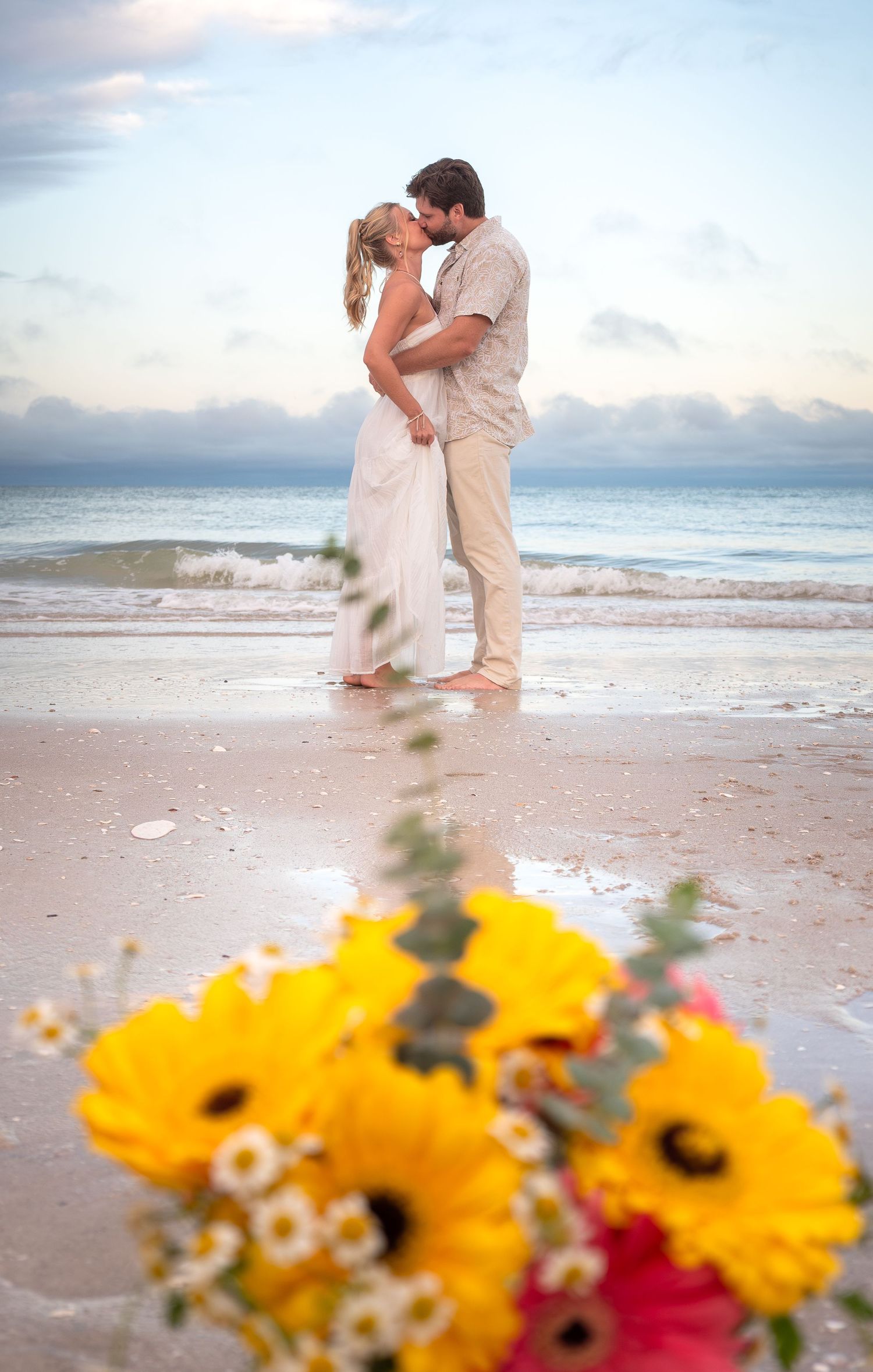 Couple kissing on beach at sunset with yellow flowers in foreground during romantic beach wedding ceremony.