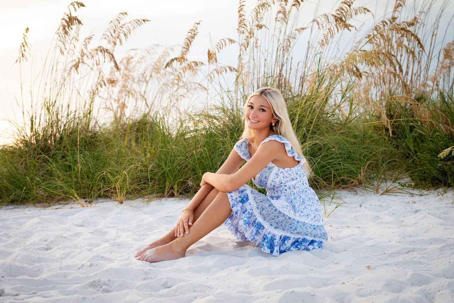 Blonde woman in blue floral dress sitting on white sand beach with sea oats and green dunes in background.