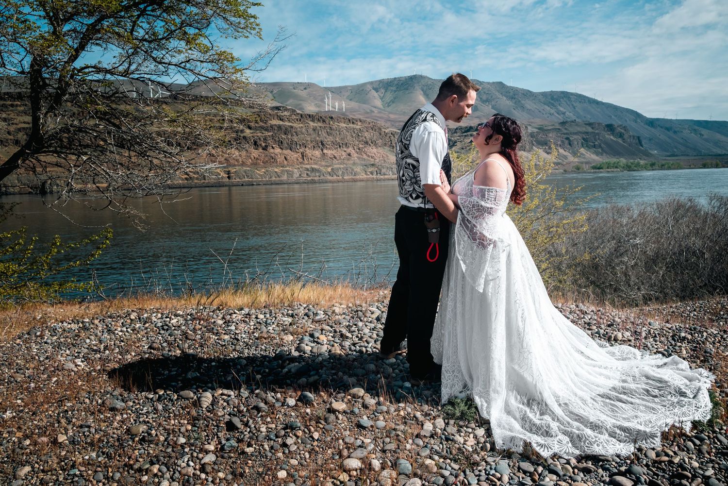 Bride and groom share a romantic moment by a mountain lake, bride in flowing white gown on rocky shore.