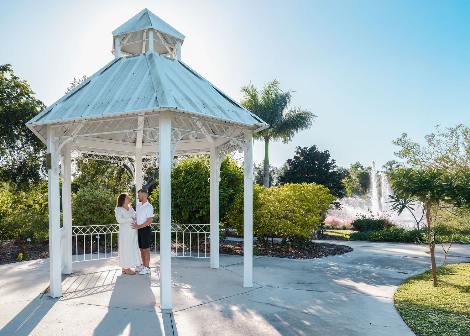 Couple embracing under a white gazebo surrounded by lush greenery and a fountain in background.