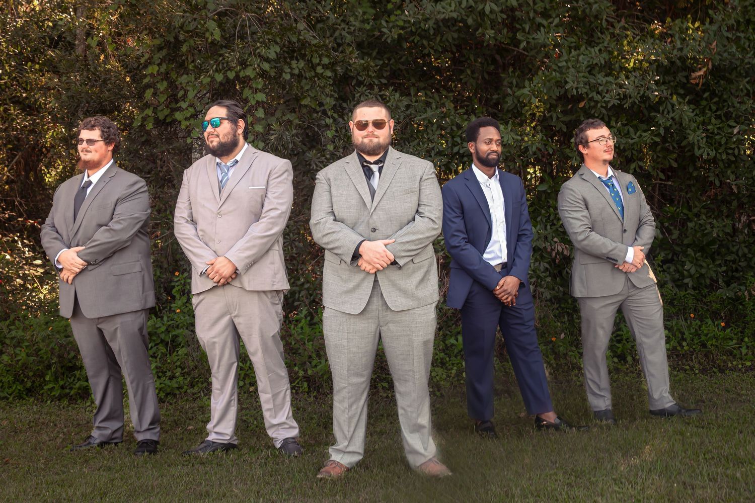 Five groomsmen in gray and navy suits posing outdoors on grass at a wedding.