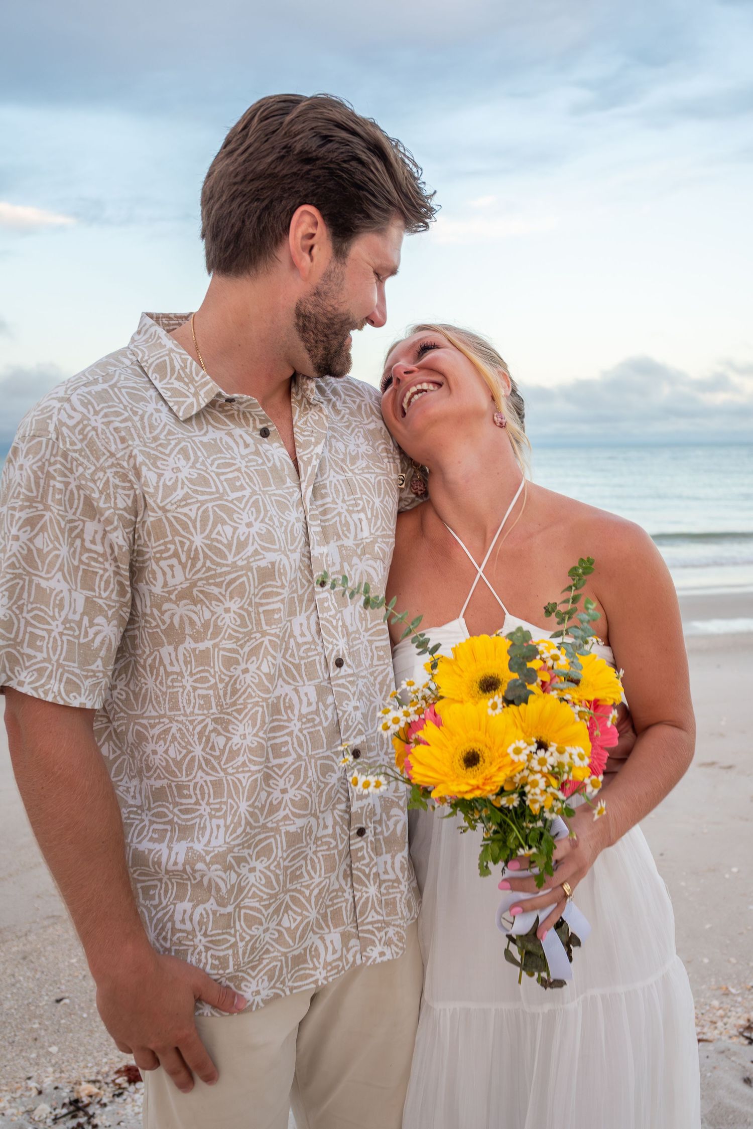 Couple sharing a romantic moment on the beach, woman holding colorful yellow and pink flower bouquet in white dress.