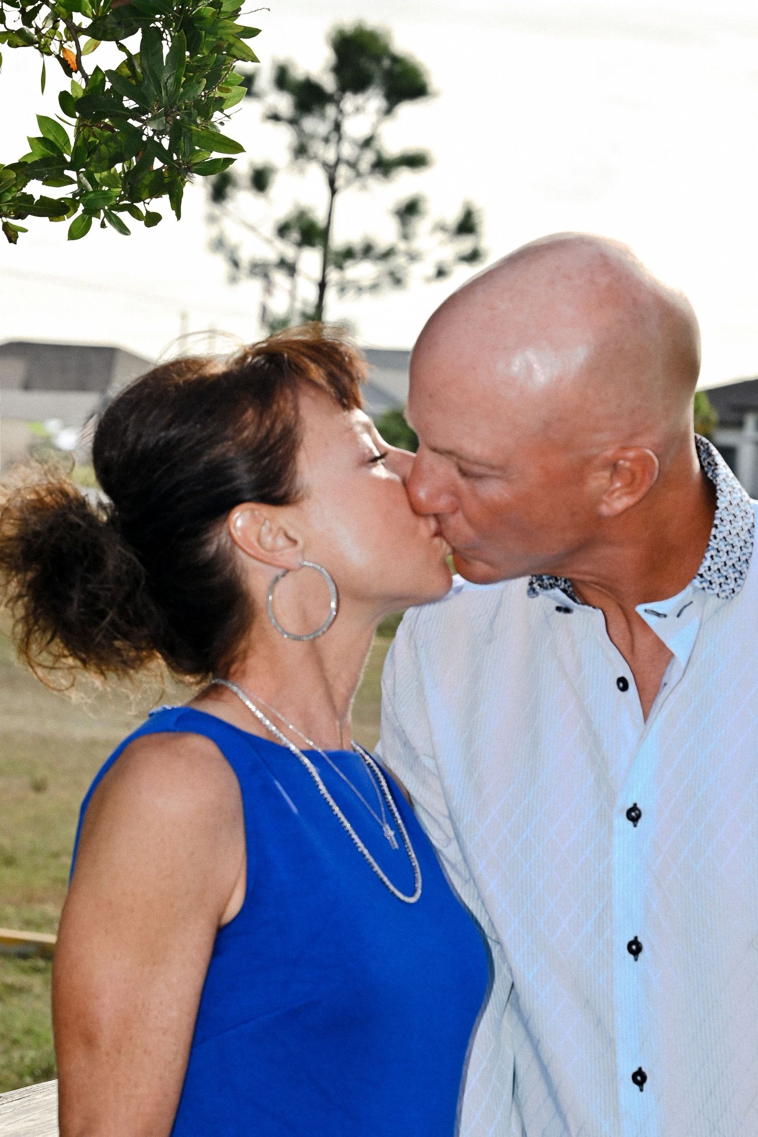 Mature couple sharing a romantic kiss outdoors, woman in blue dress wearing hoop earrings.