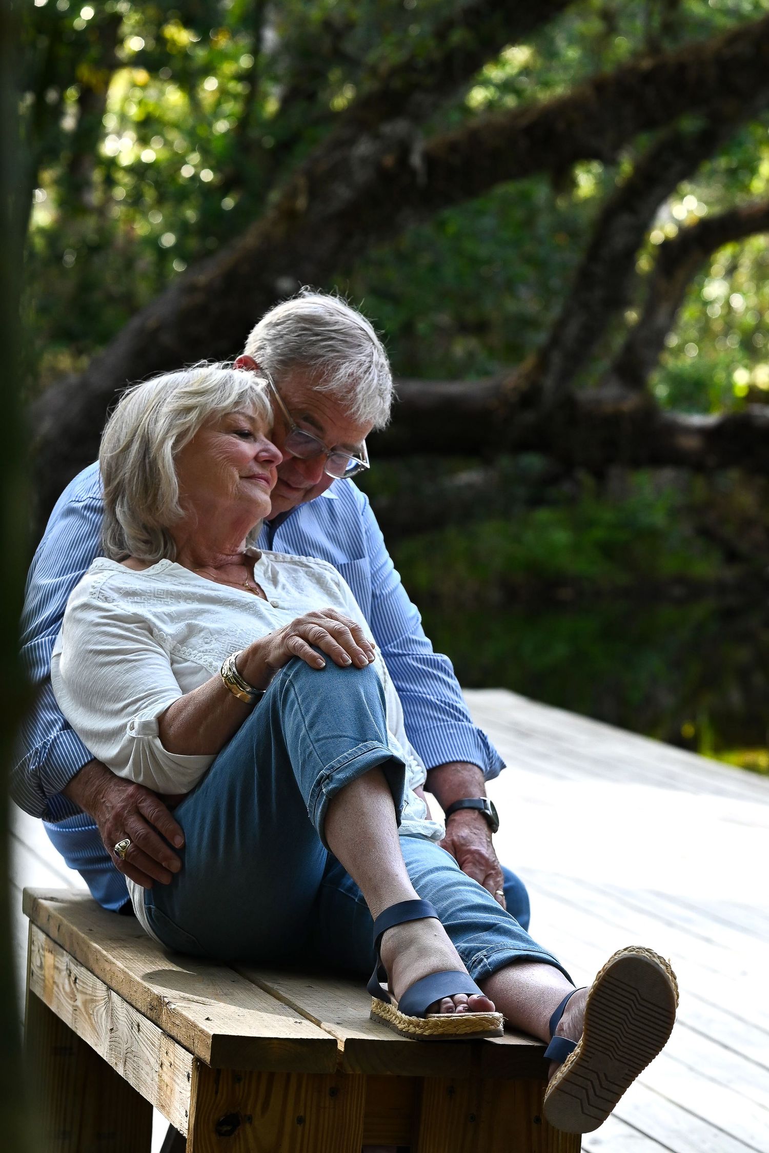Senior couple sitting closely on wooden deck surrounded by lush green trees, sharing tender moment.