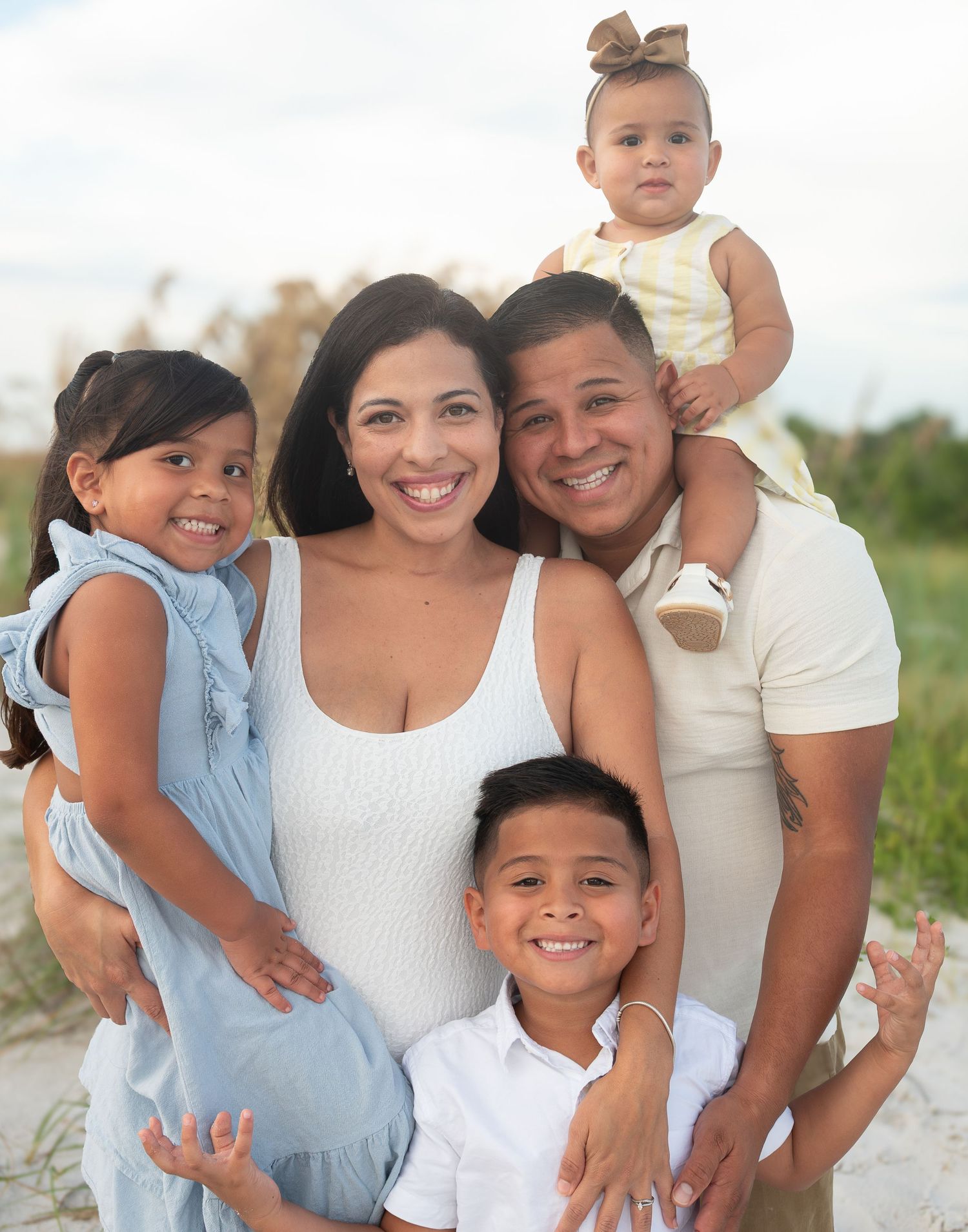A smiling Hispanic family of five poses together on a sandy beach, including three young children.