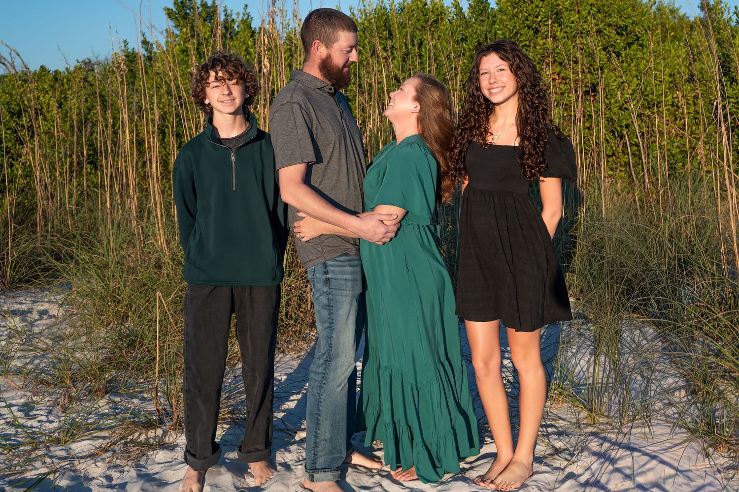 A family of four stands together on sandy dunes near tall green beach grass.