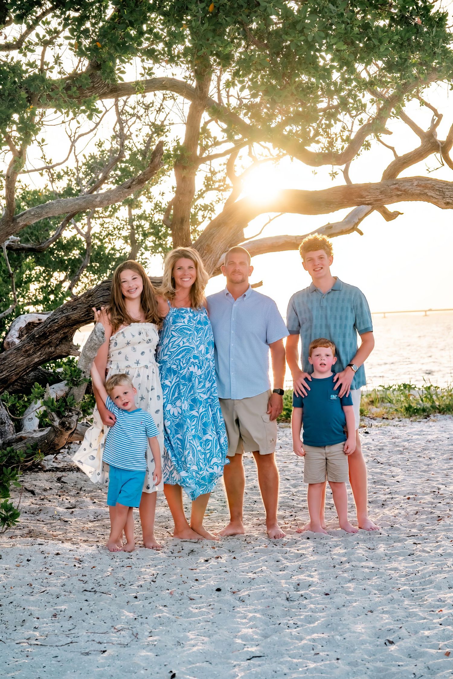 Family of six posing on a sandy beach at golden hour beneath a sprawling tree with warm sunset light.