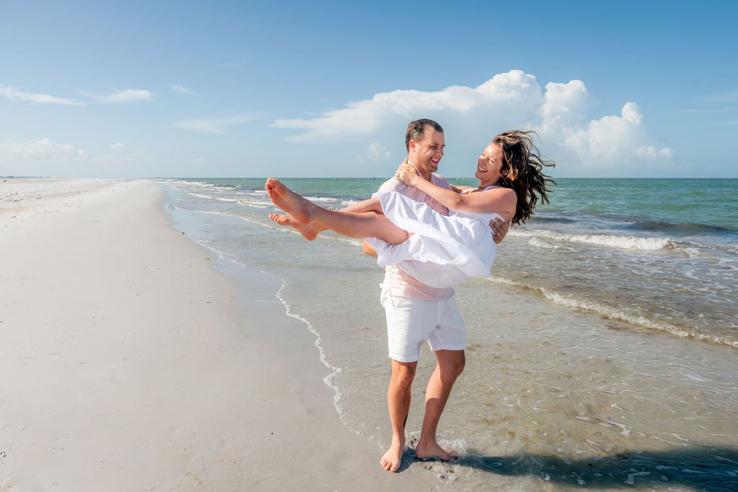 A couple in white outfits celebrating on a sunny beach, man lifting woman as waves wash ashore.