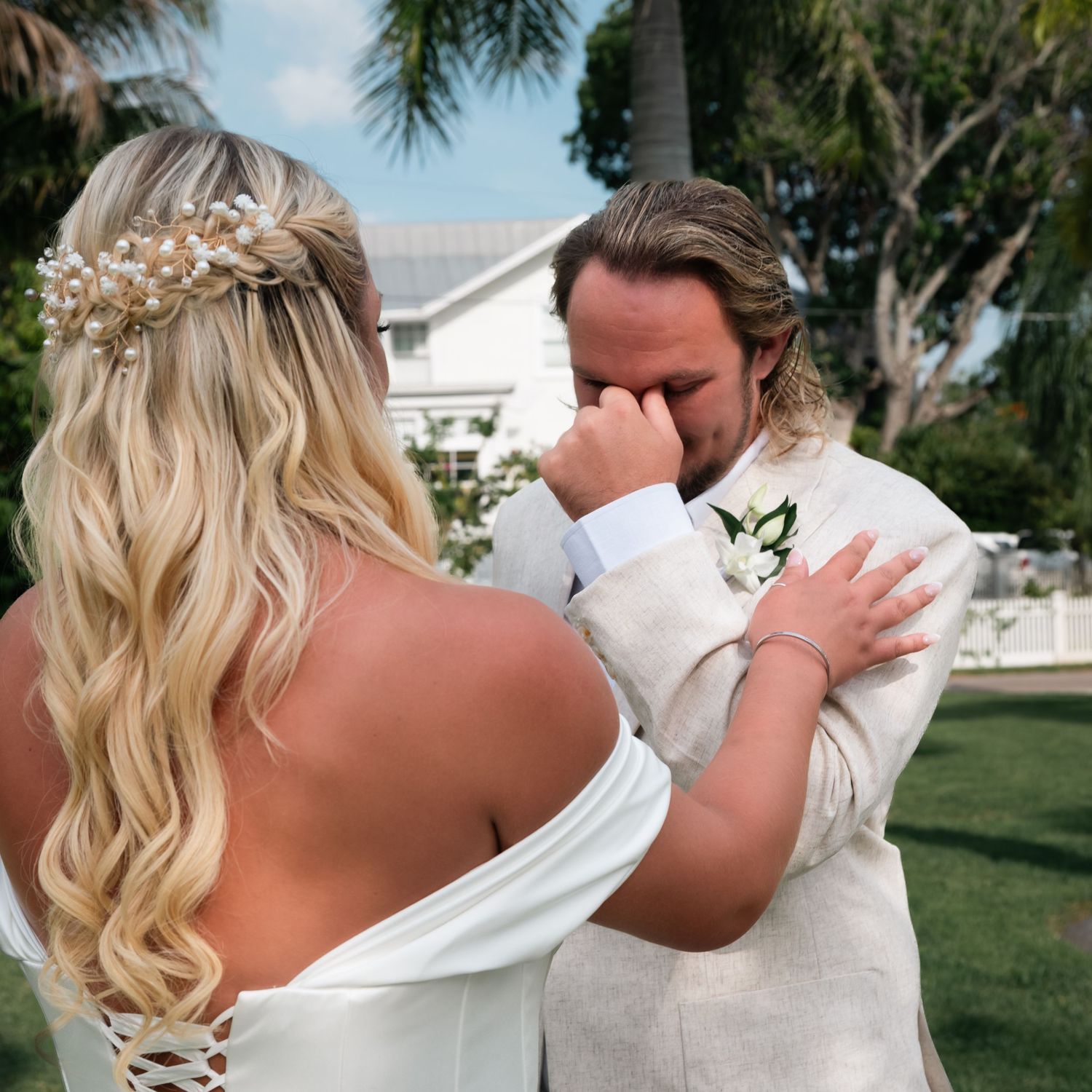 Emotional groom wiping tears during first look with bride in white off-shoulder gown and pearl hair accessory.