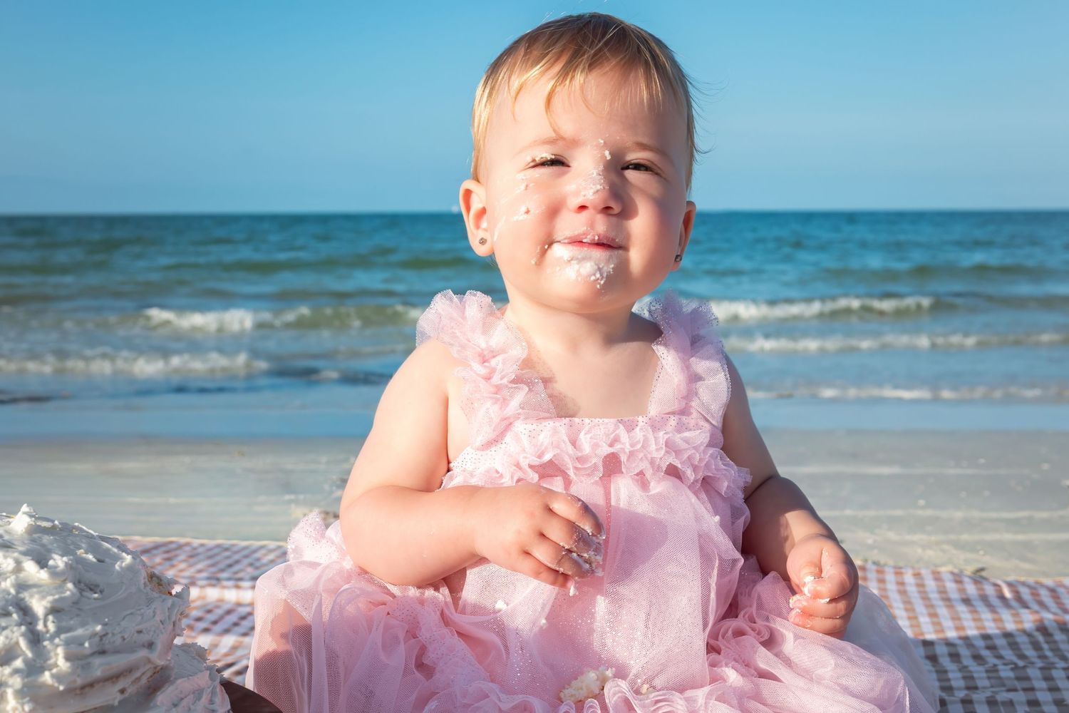A happy baby girl in a pink ruffled dress sits on a beach with ocean waves in the background.