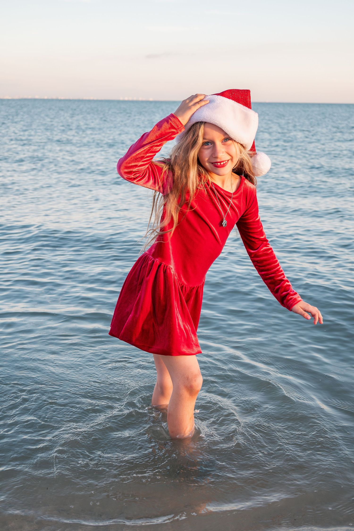 Young girl in red velvet dress and Santa hat playing in ocean waves at sunset.