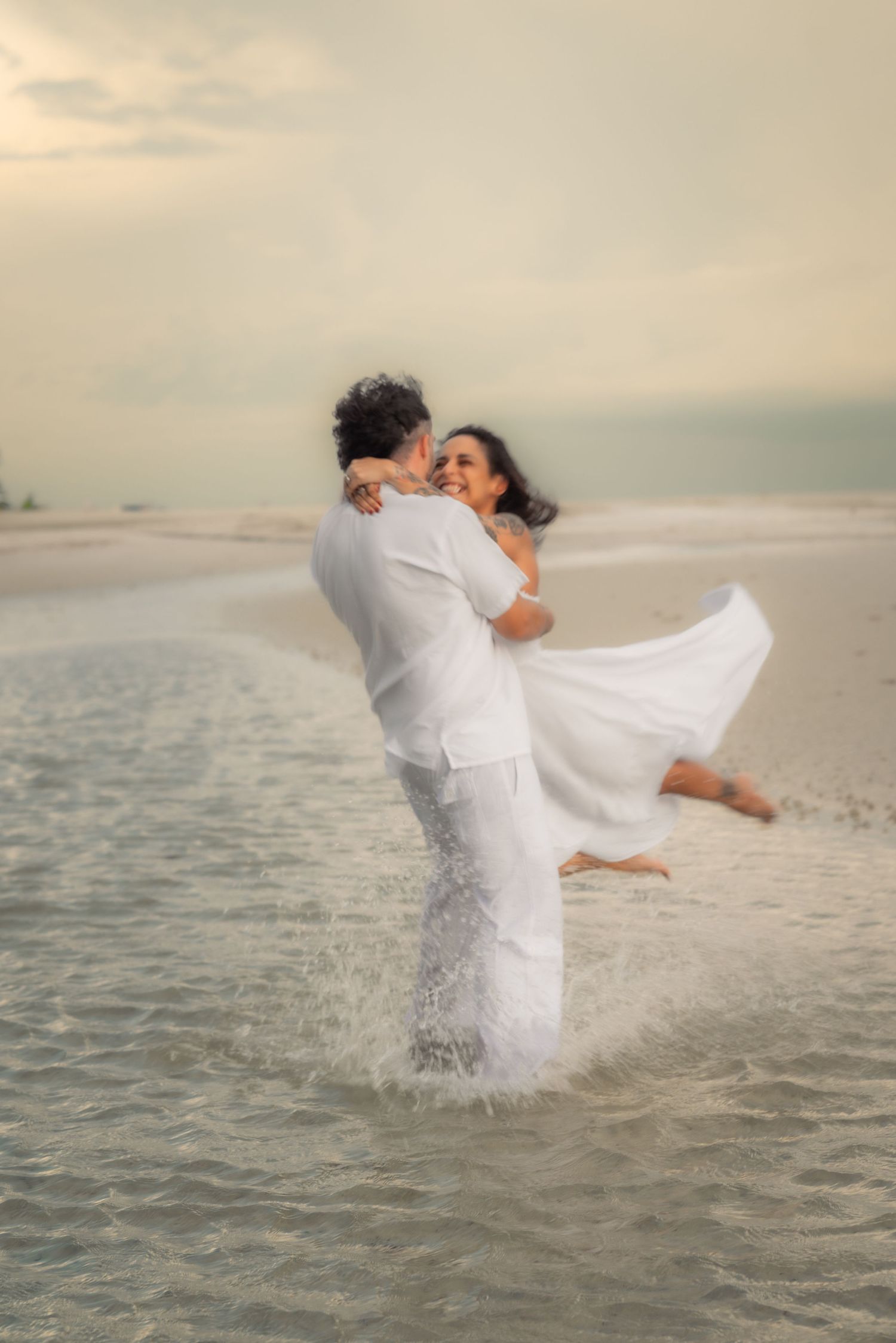 Man in white lifting smiling woman in white dress while standing in ocean waves on beach.