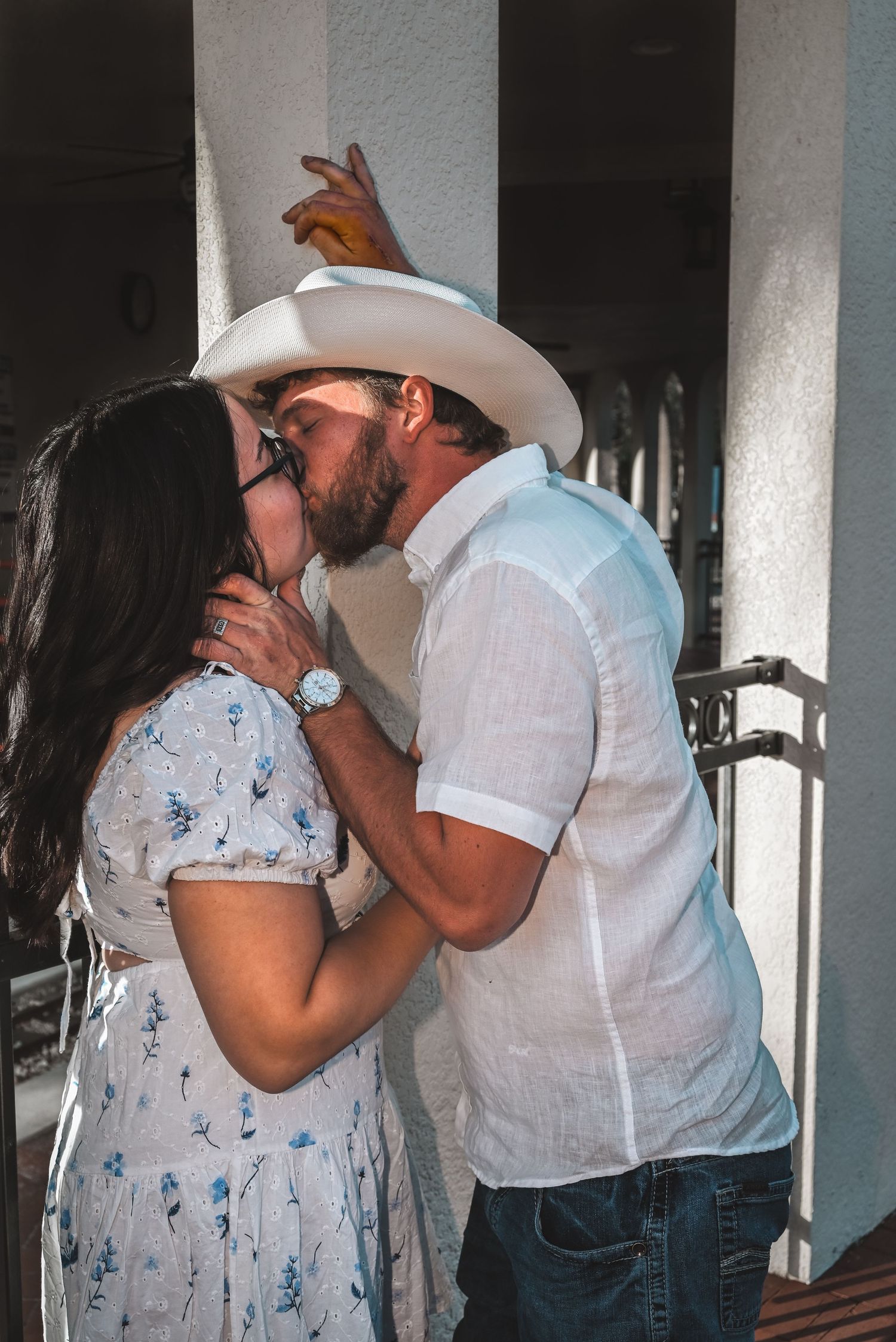 Couple sharing a romantic kiss, man wearing white cowboy hat and linen shirt outdoors.