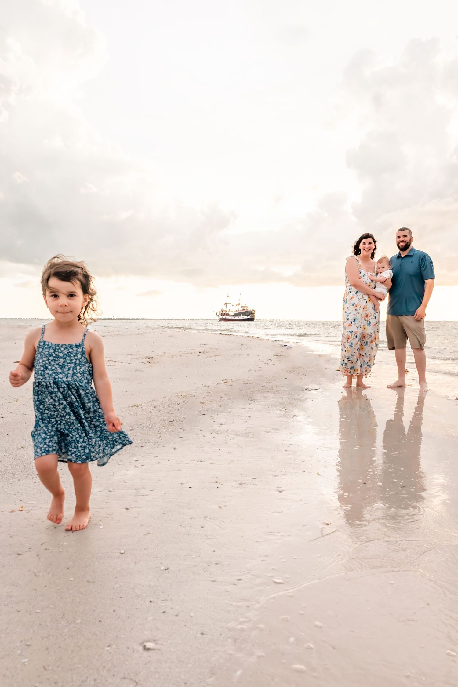 Family beach portrait with toddler girl in blue dress running on sandy shore while parents hold baby at sunset.