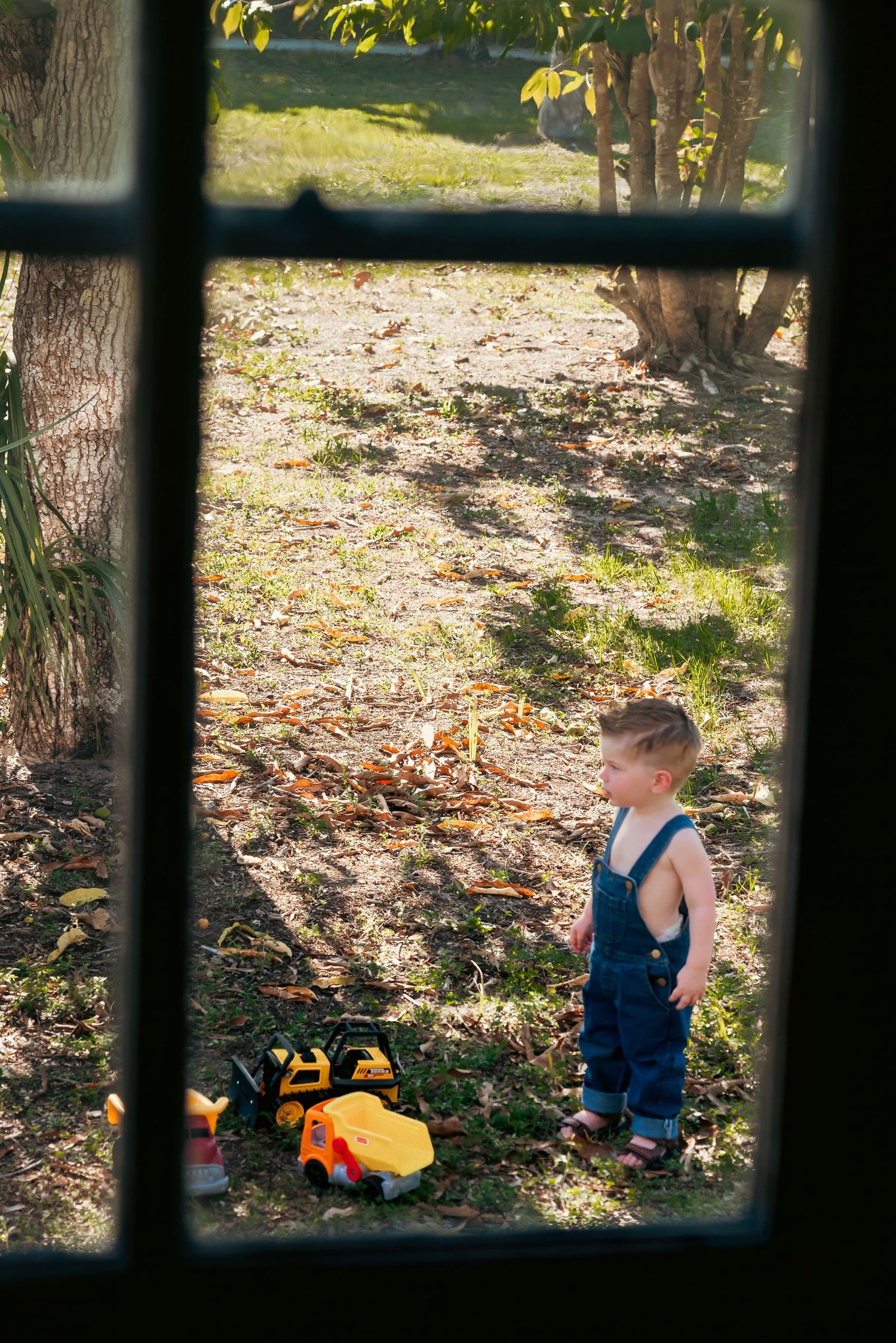 Young toddler boy in denim overalls playing with toy trucks outdoors, viewed through a window frame.