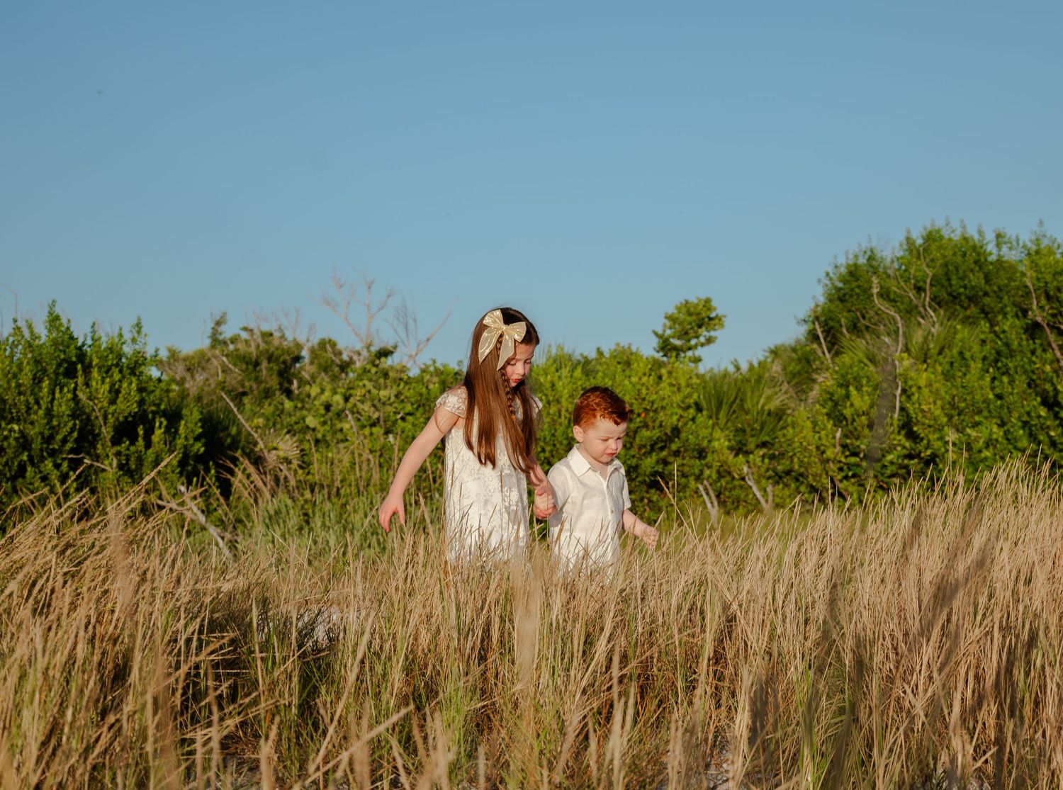 Young girl and redhead boy walking together through tall golden grass in a sunny natural landscape.