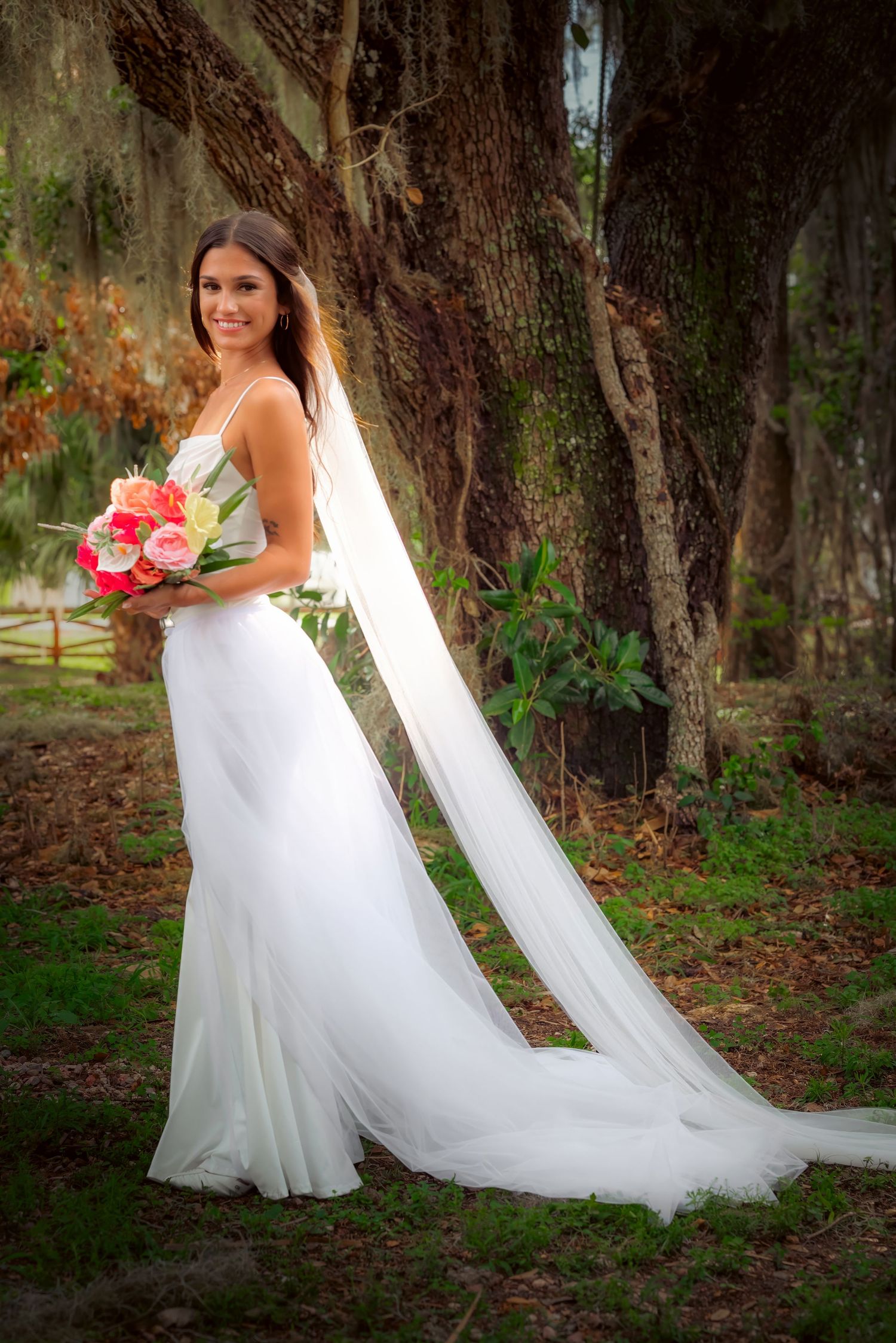 Smiling bride in elegant white gown with long veil holds a pink bouquet beside a large oak tree.