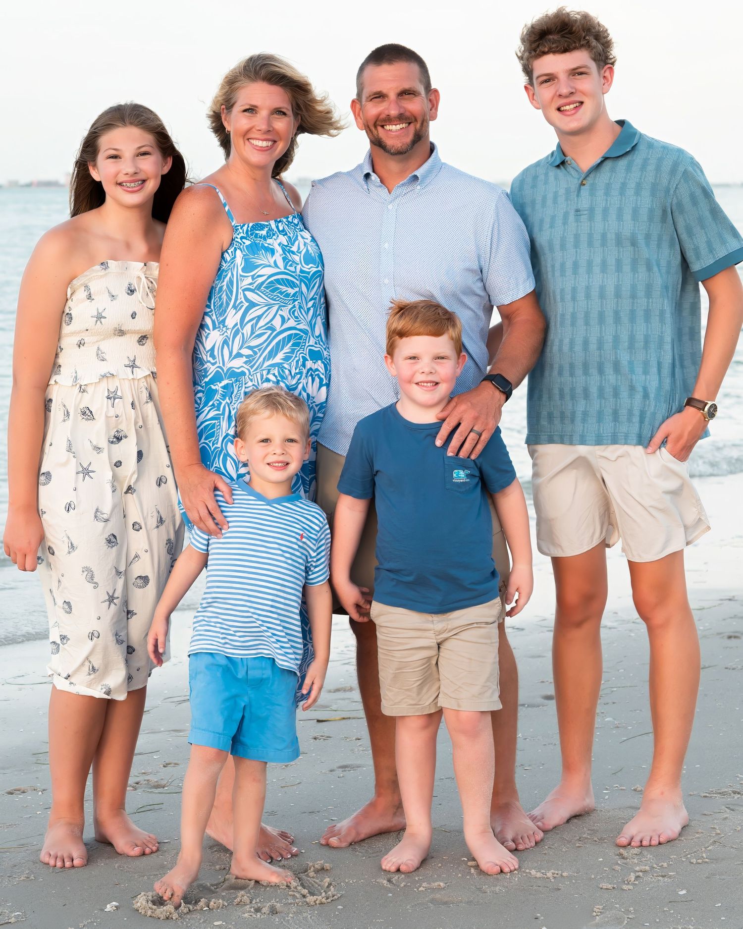 Happy family of six posing together on a sandy beach, smiling in coordinating blue and white outfits.