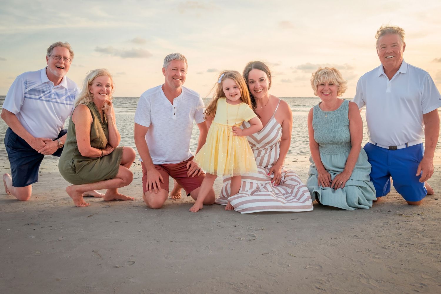 Multi-generational family of seven smiling together on a sandy beach at sunset in casual attire.