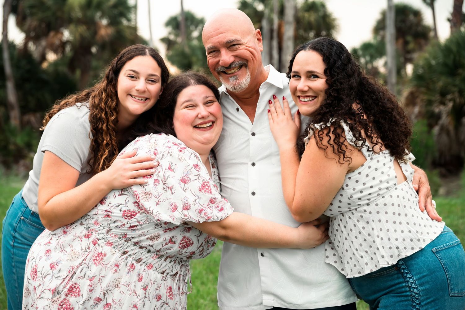 A smiling family of four poses outdoors, with a bald man and three women with curly hair hugging closely.