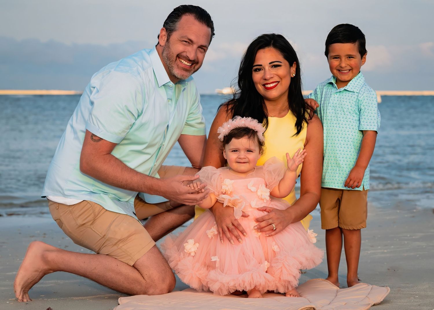 Family of four posing on beach at sunset, parents with toddler girl in pink dress and young boy in teal shirt.
