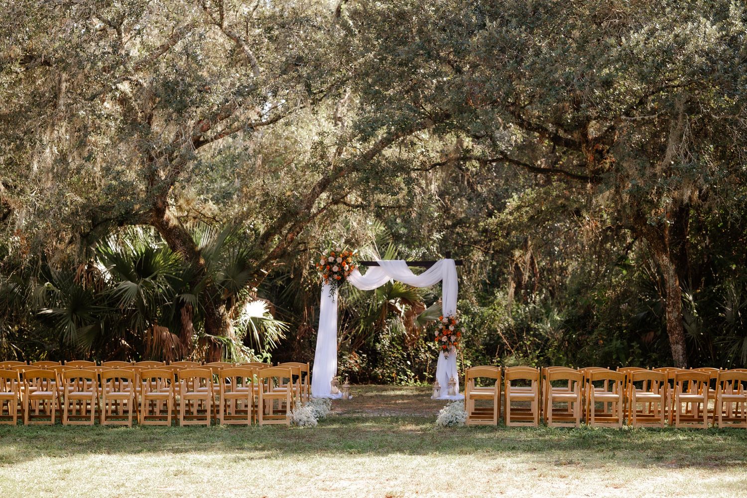 Outdoor wedding ceremony setup with wooden chairs, white floral arch, and lush oak tree canopy in Florida.