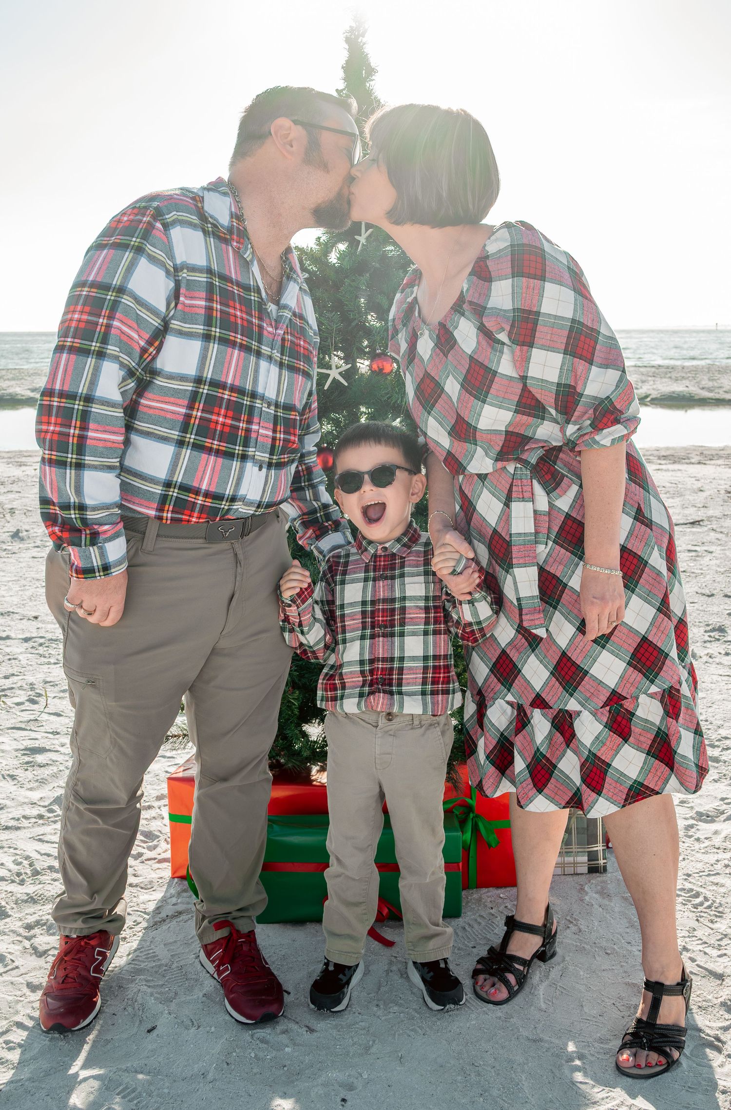Parents in matching plaid kiss over surprised young boy in sunglasses during fun Christmas beach family photo session.