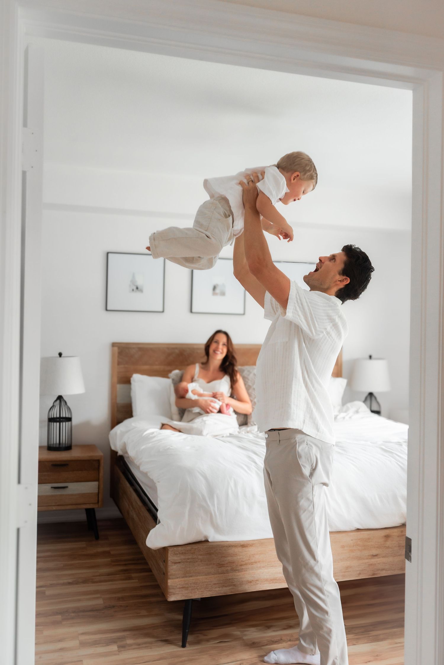 Father lifting toddler in air while mother holds newborn in bedroom.