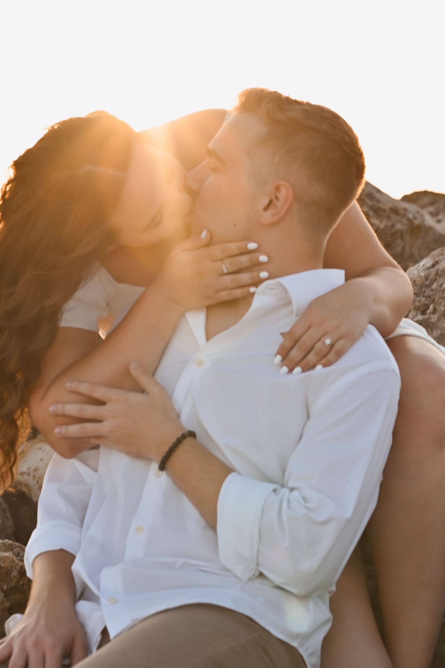 Couple kissing passionately while sitting on rocks bathed in warm golden sunset light.