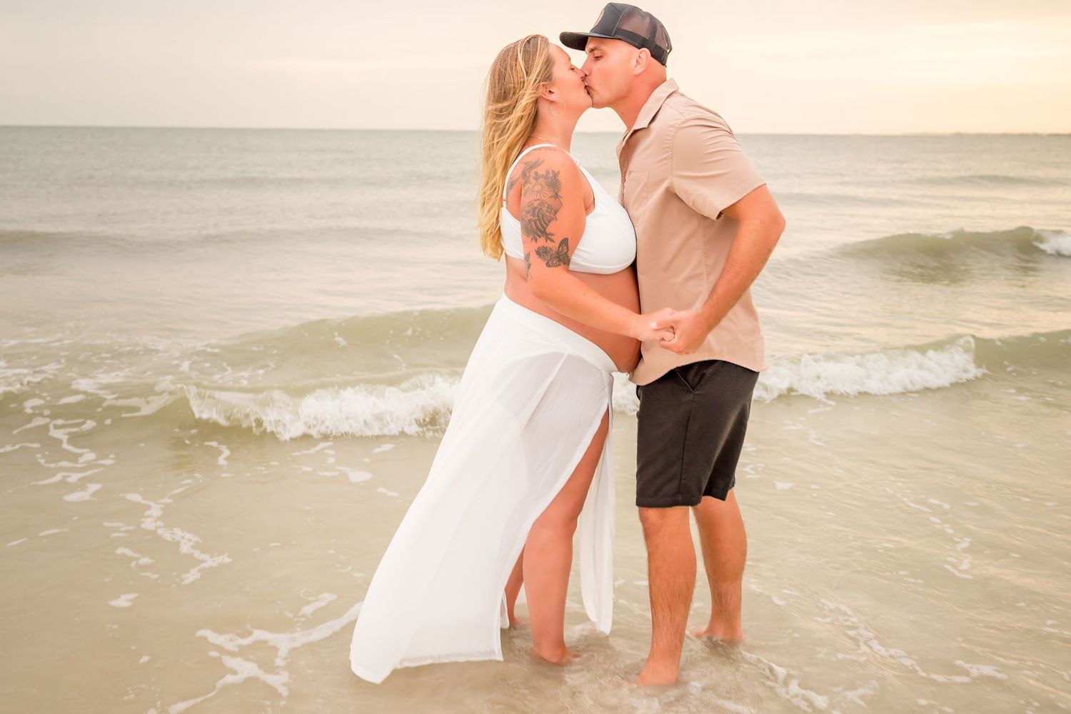 Couple kissing on a sandy beach at sunset, woman in white dress, man in khaki shirt and black shorts, ocean waves behind them.