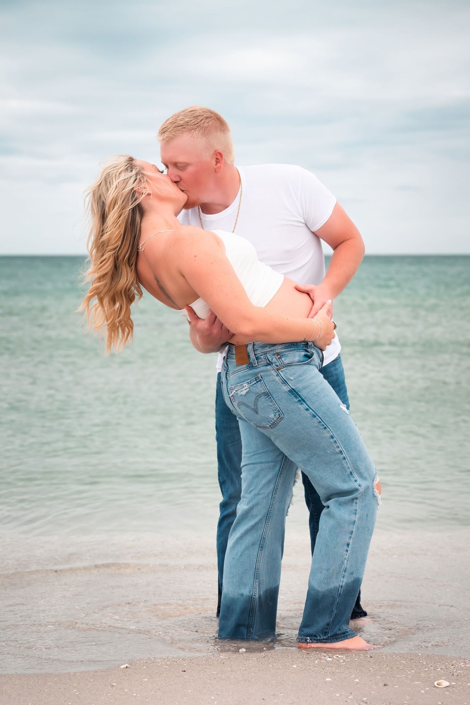 Couple sharing a romantic kiss on the beach, dressed in white tops and jeans, with ocean waves in the background.