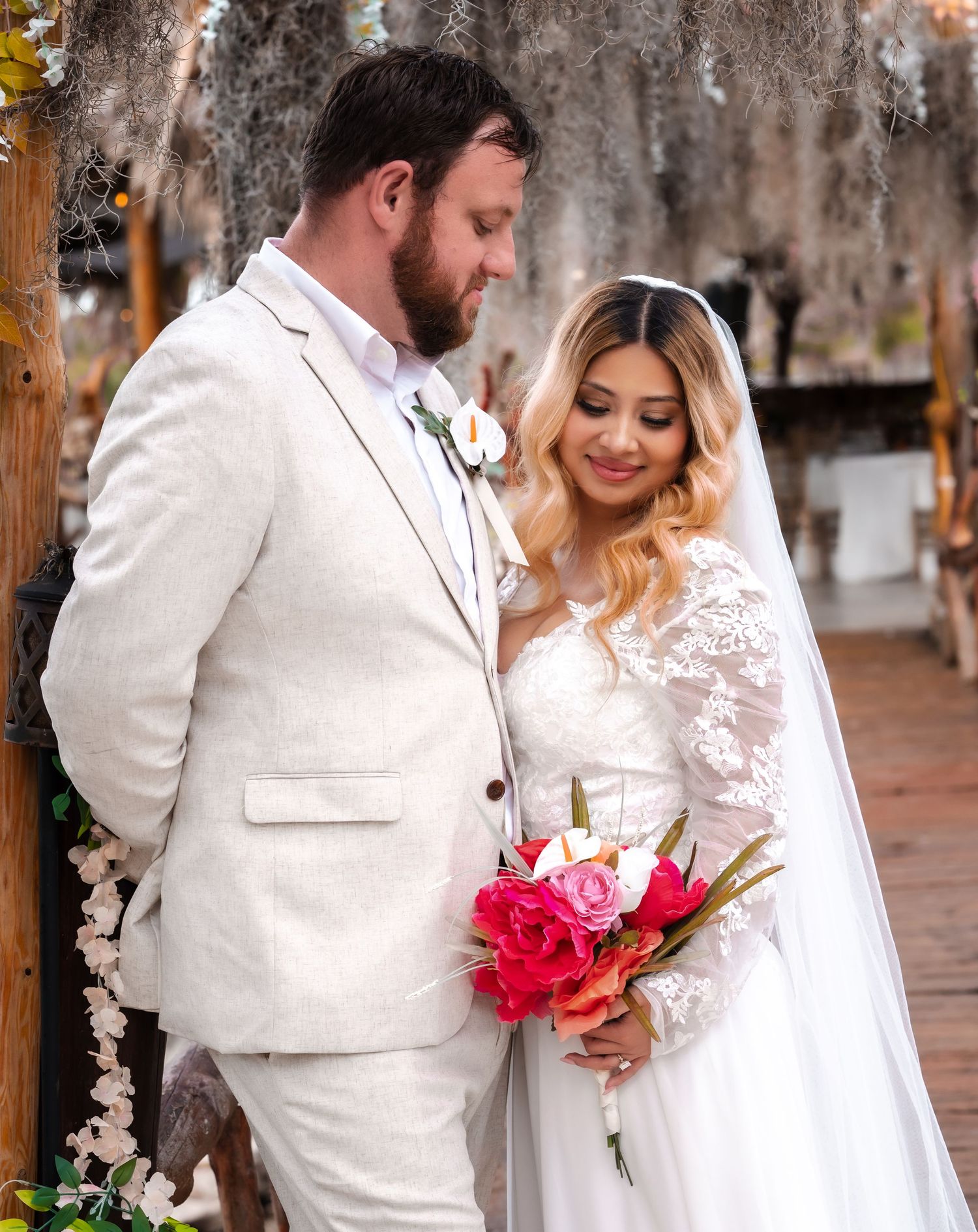 Bride in lace gown holding pink bouquet stands with groom in white suit during outdoor wedding ceremony.