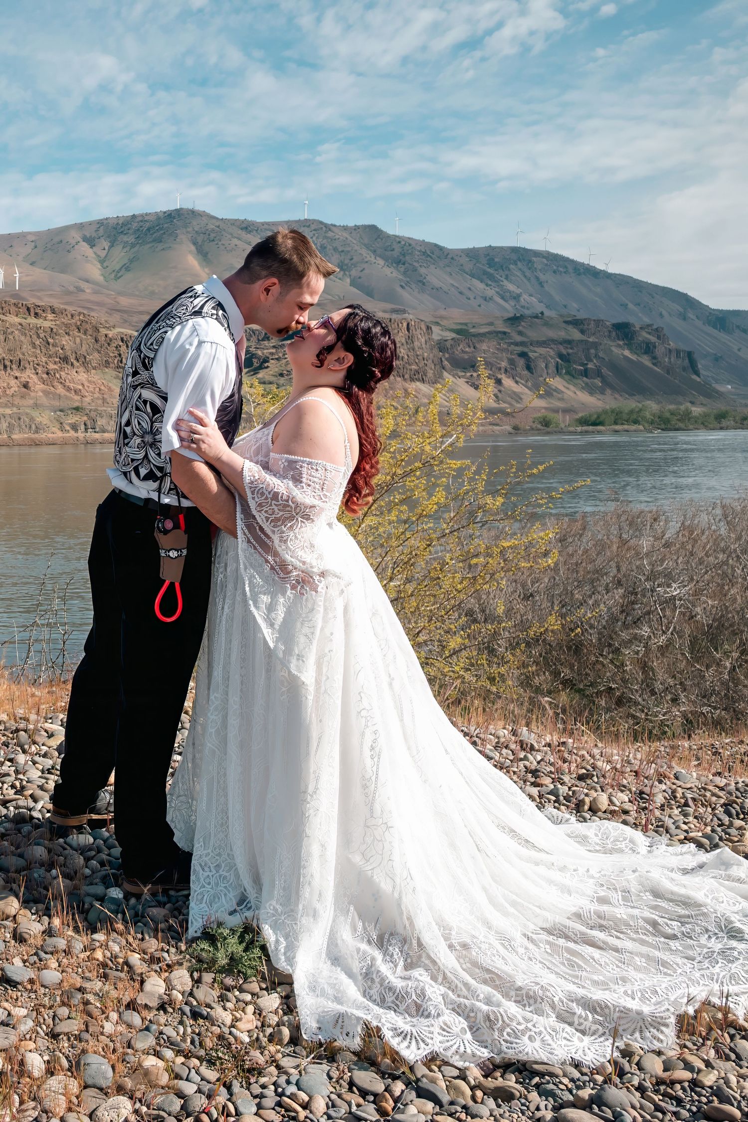 Bride and groom share romantic moment by mountain river, bride in flowing lace gown, groom in vest on rocky shore.