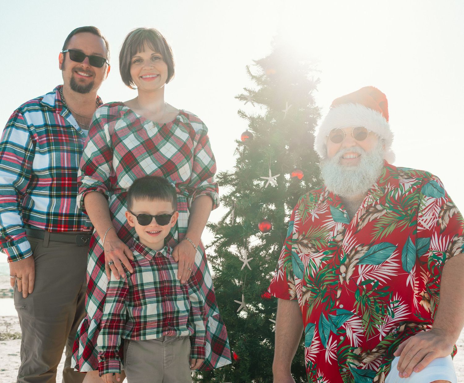 Family of four poses with beach Santa Claus beside Christmas tree on sunny Florida beach holiday photo session.